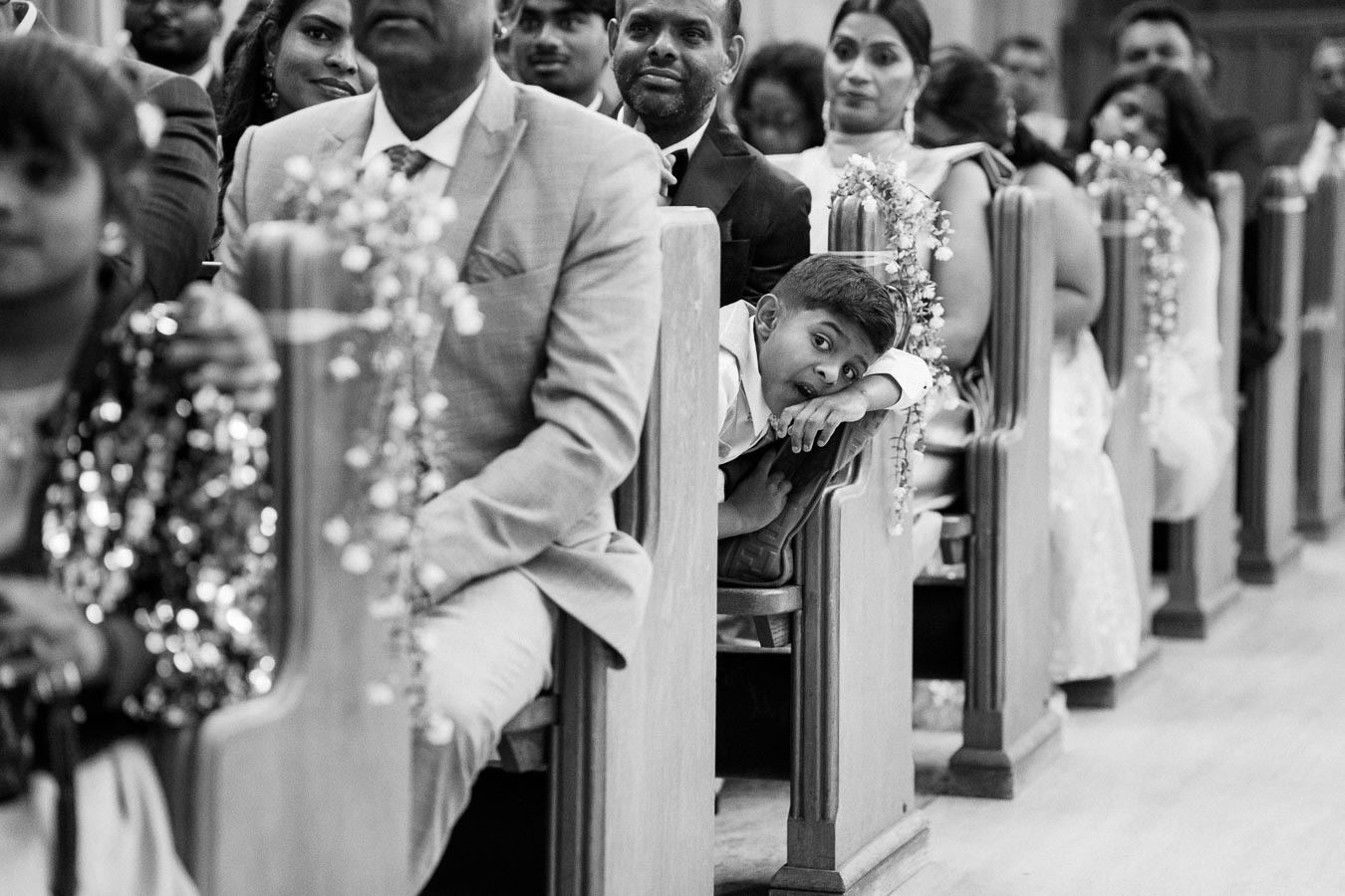 Black and white image of a wedding ceremony with guests seated in decorated pews, featuring a young boy playfully leaning over, capturing a joyful moment.