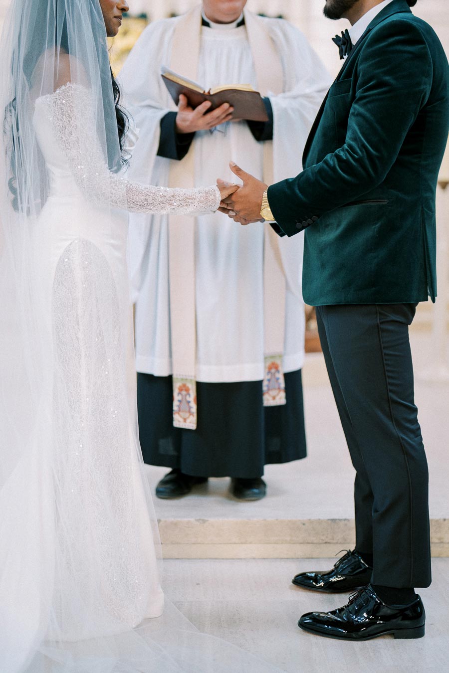 Bride and groom holding hands during a wedding ceremony in a church, with an officiant reading from a book; the bride is wearing a white gown and veil, and the groom is in a dark suit.