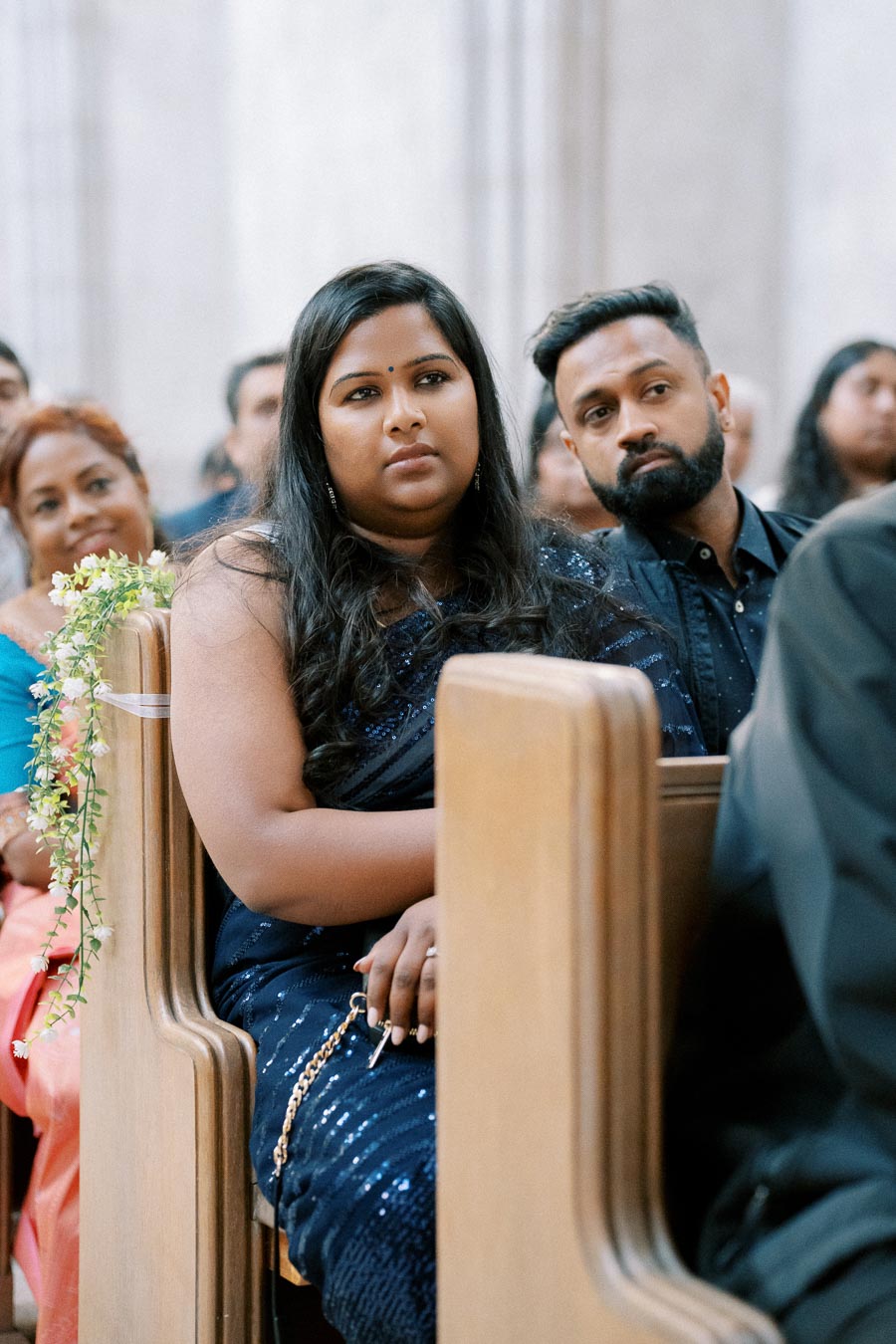 A group of people sitting in a church, attentively watching an event. The focus is on a woman in a dark blue dress sitting next to a man with a beard, both showing expressions of interest and engagement. Decorated pews and other attendees in the background suggest a ceremony or formal gathering.