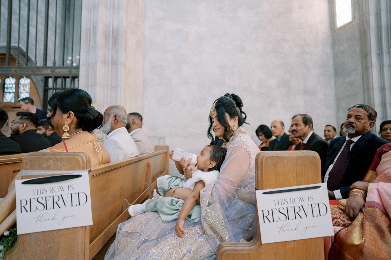 Mother feeding baby with bottle in a church reserved seating area, surrounded by elegantly dressed guests during a ceremony.