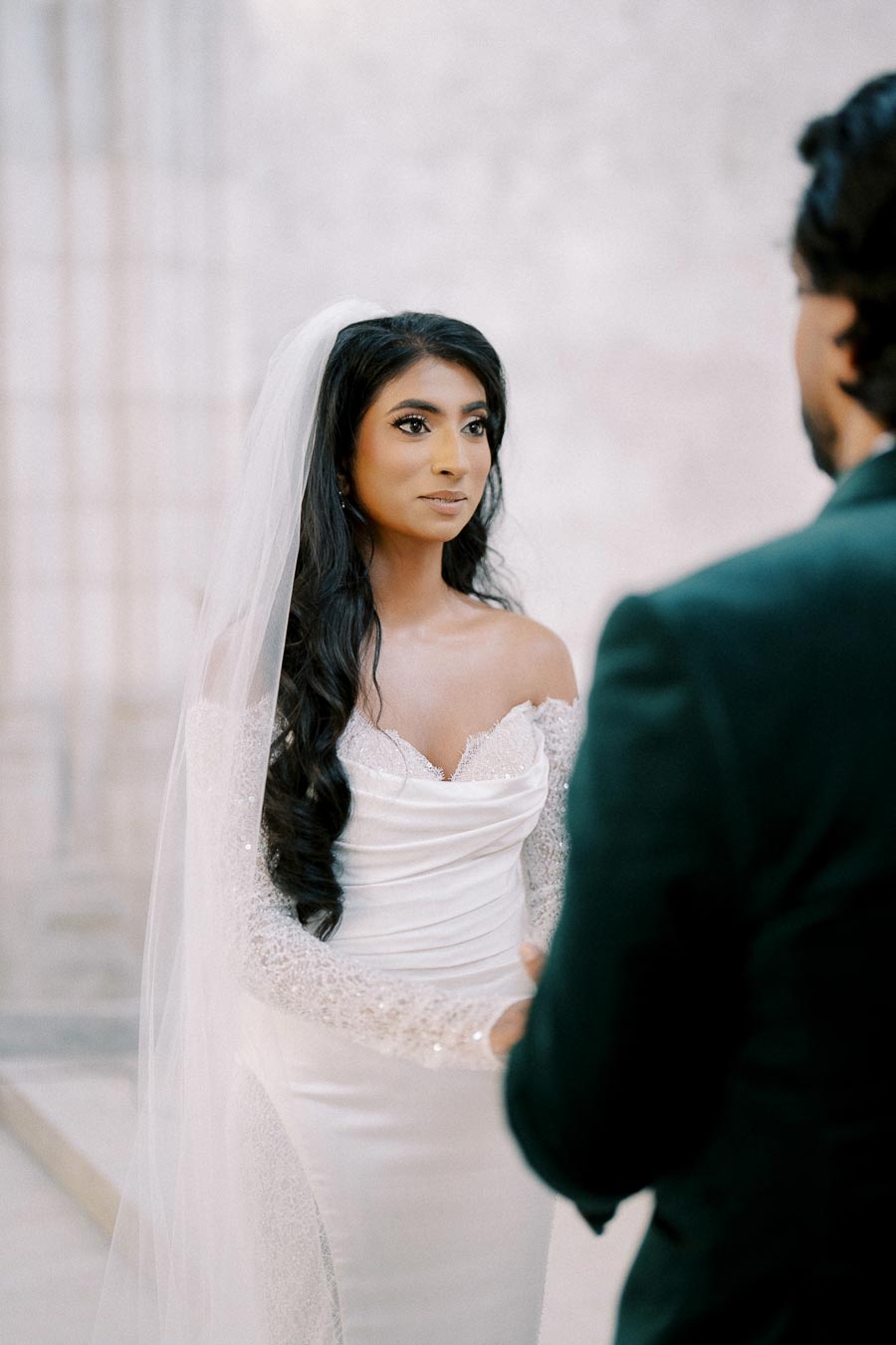 Bride in an elegant lace wedding dress and veil, standing in front of groom during a ceremony, captured in soft lighting.