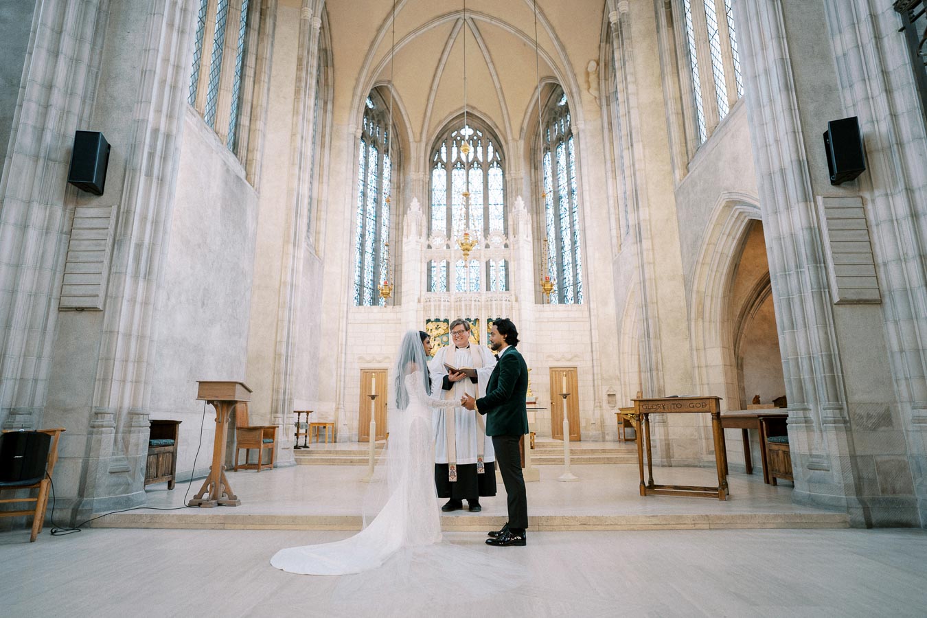 Bride and groom exchanging vows in a beautiful, ornate church with high arched ceilings and stained glass windows, attended by a priest.