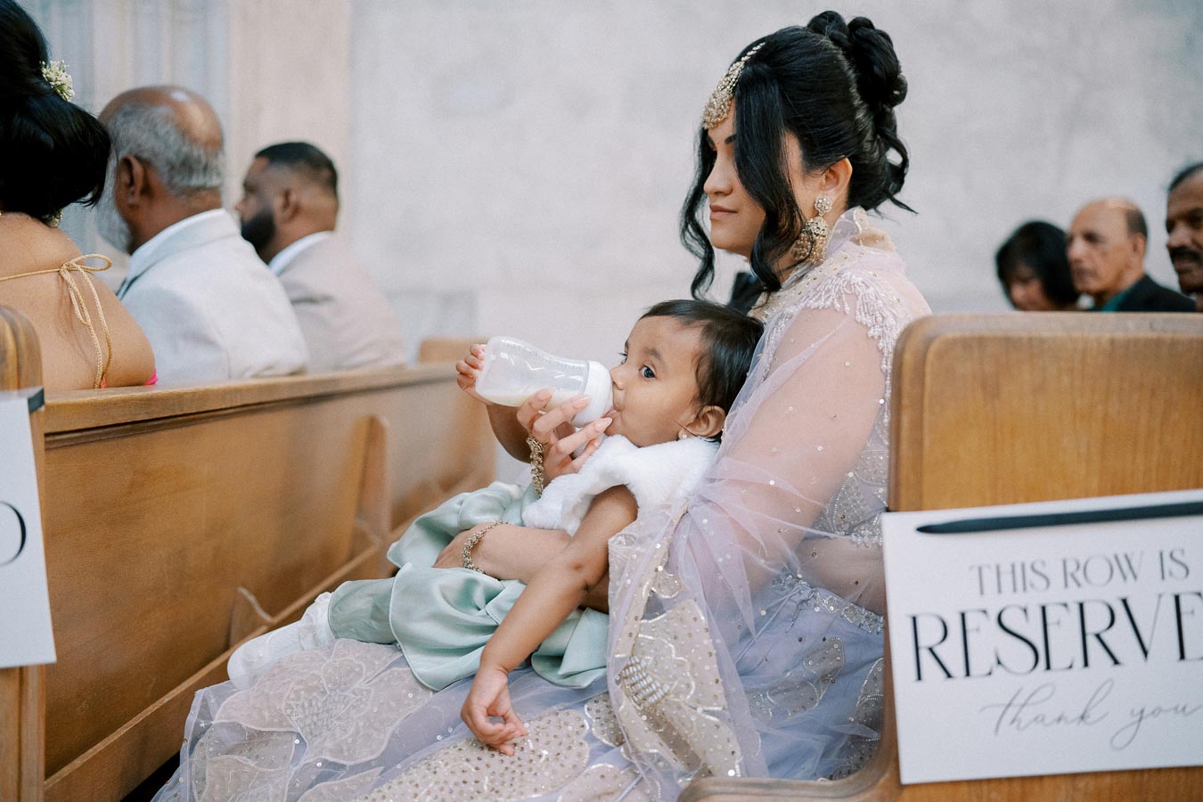A woman dressed in elegant traditional attire feeds a baby with a bottle while seated in a reserved row at a formal event.