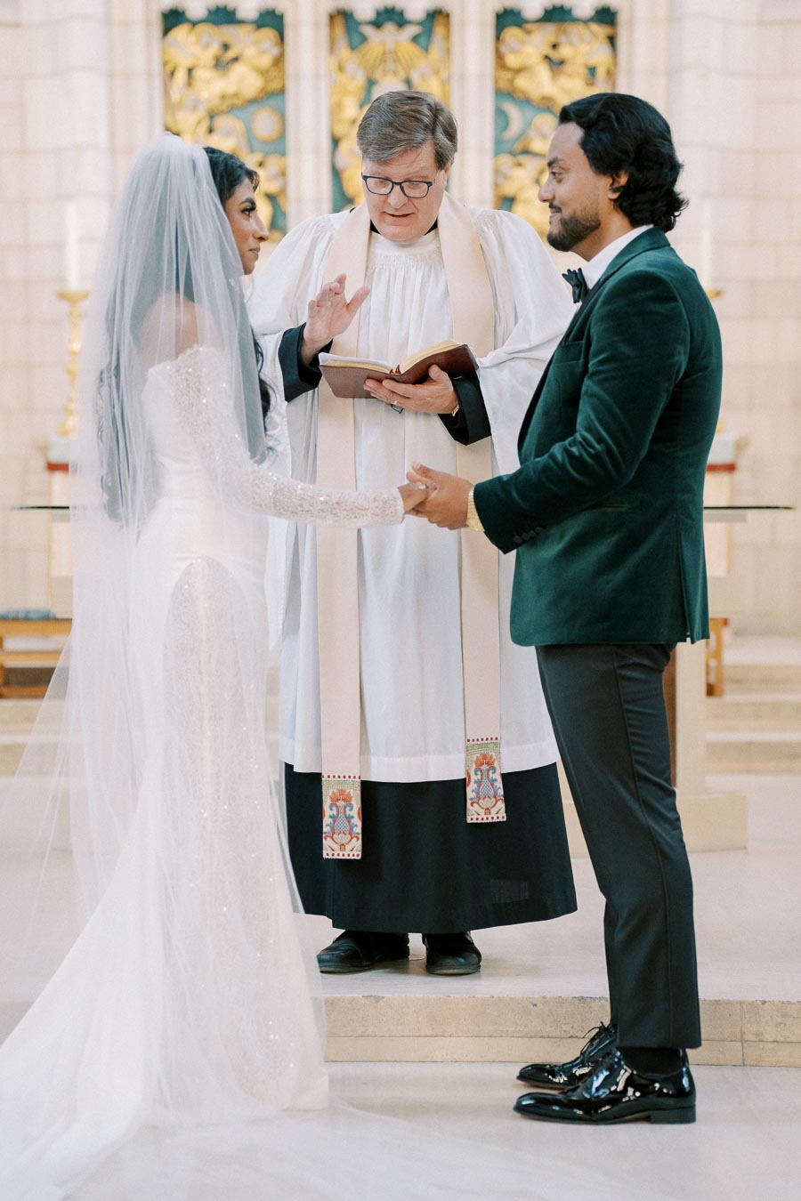 A bride in a white gown and veil holds hands with a groom in a green suit during a wedding ceremony at a church altar, officiated by a priest in robes.