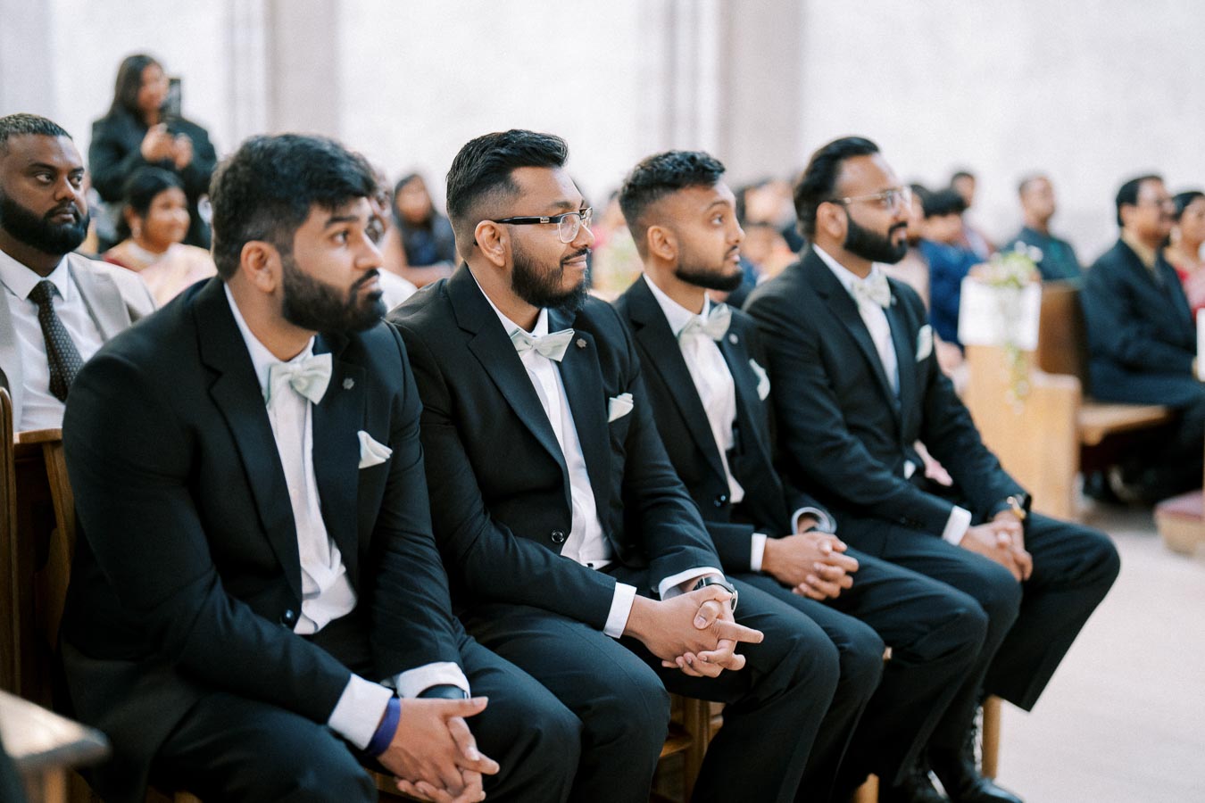 Group of groomsmen in matching black suits with bow ties sitting attentively in a church setting.