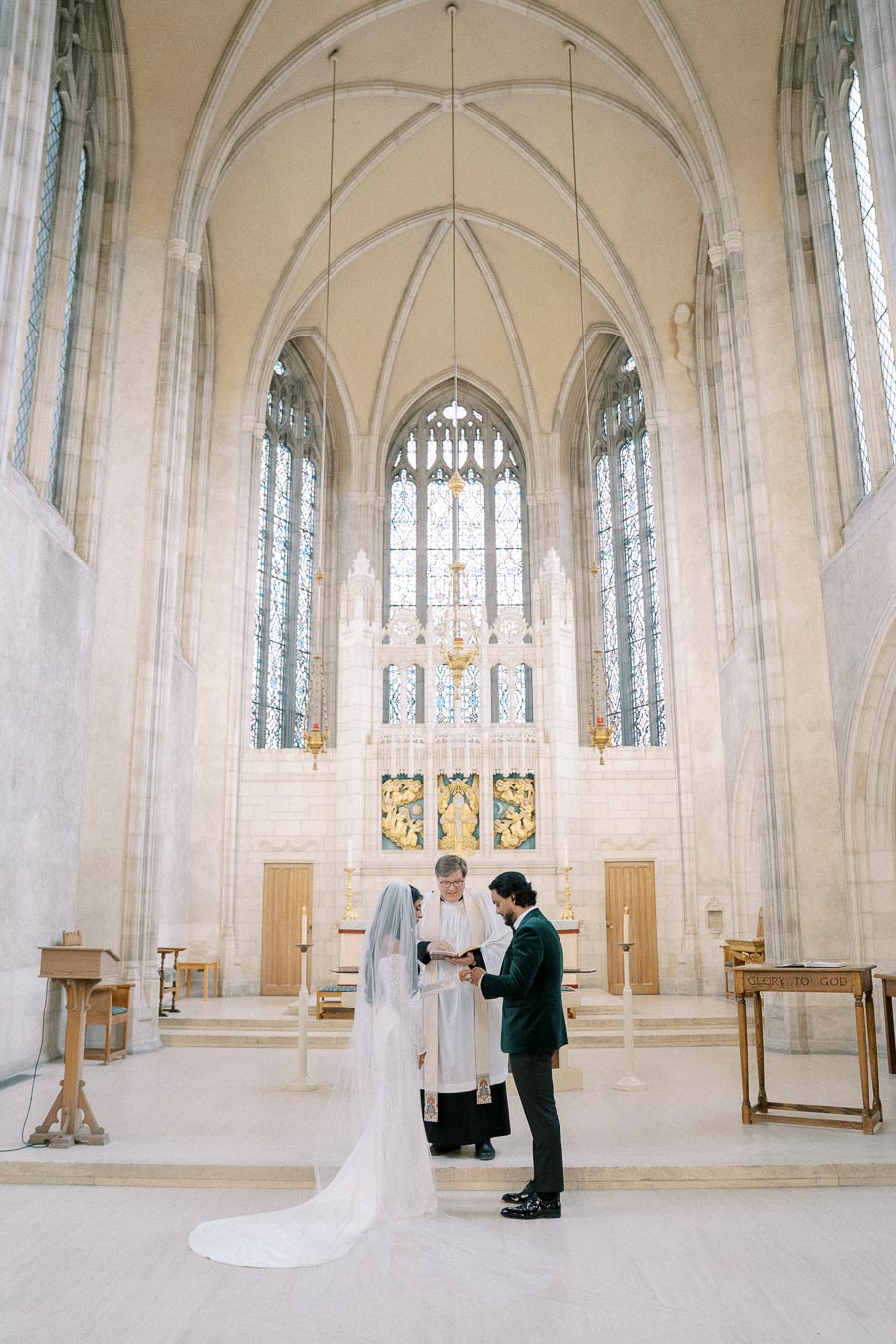 A couple exchanging vows during their wedding ceremony in a grand, historic church with high ceilings and stained glass windows.