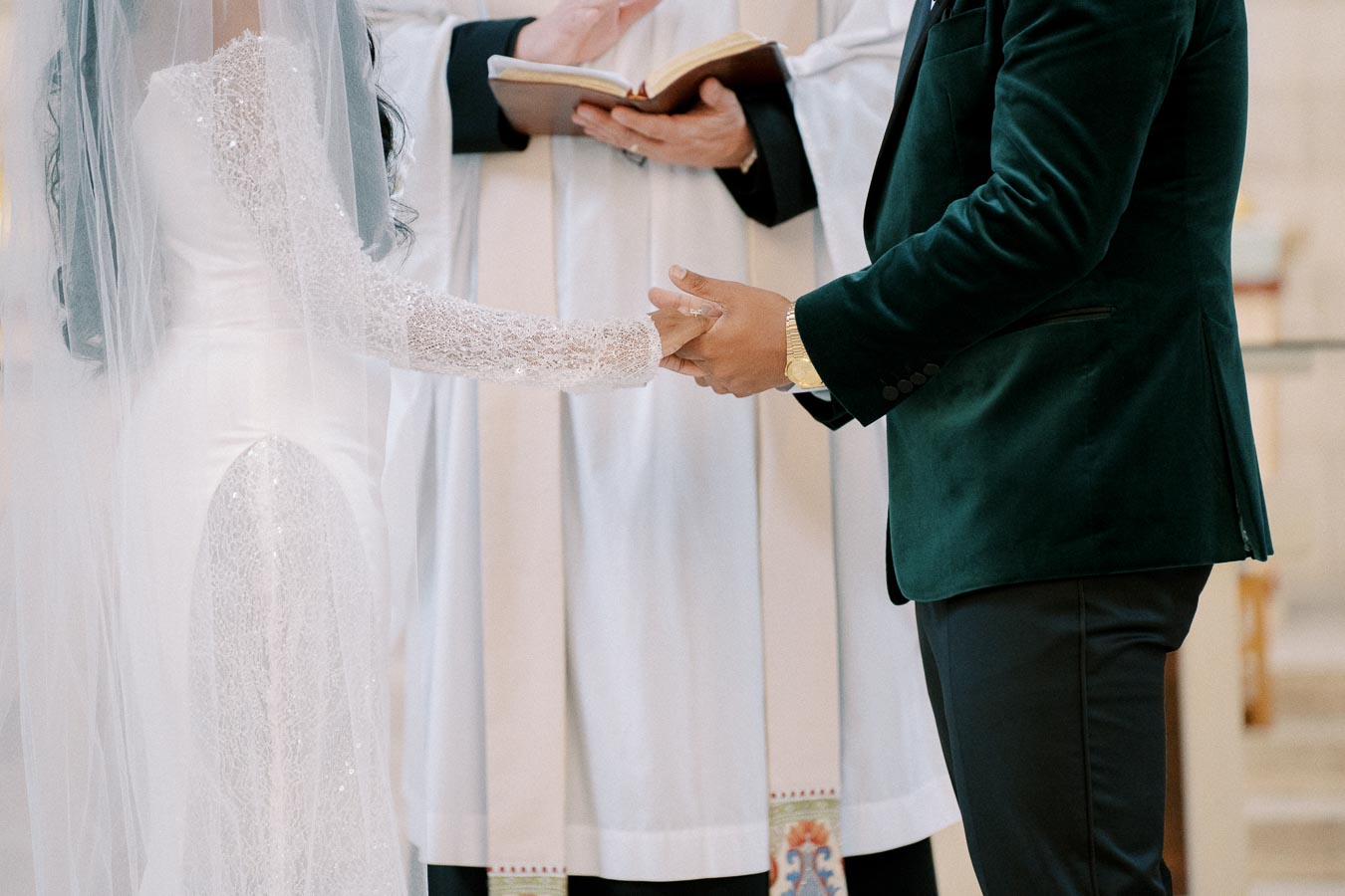 A bride and groom holding hands during a wedding ceremony, with a priest in the background holding a book, in an elegant setting.