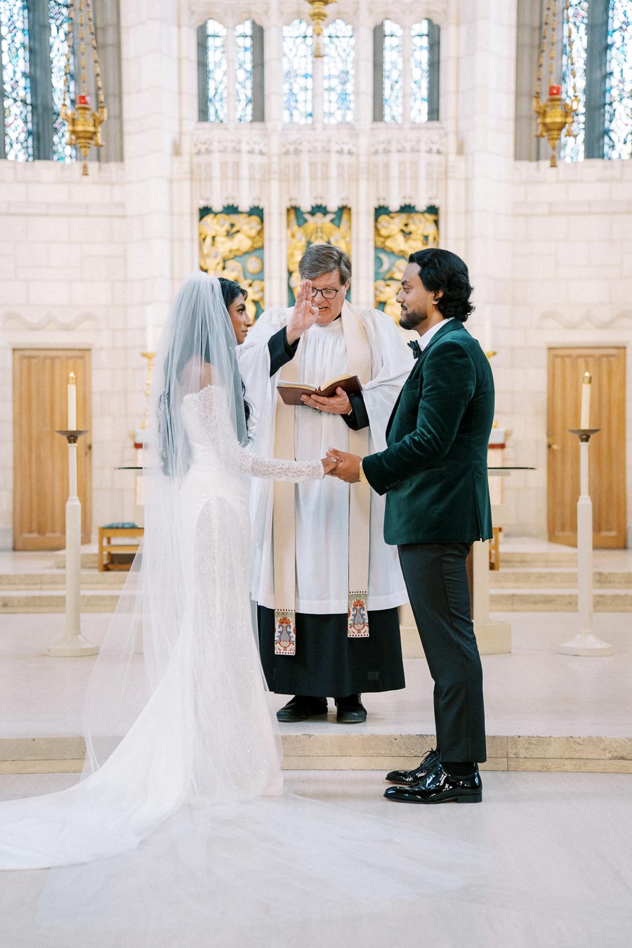 A couple exchanging vows during a wedding ceremony in a church, with a priest officiating and elegant decorations in the background.
