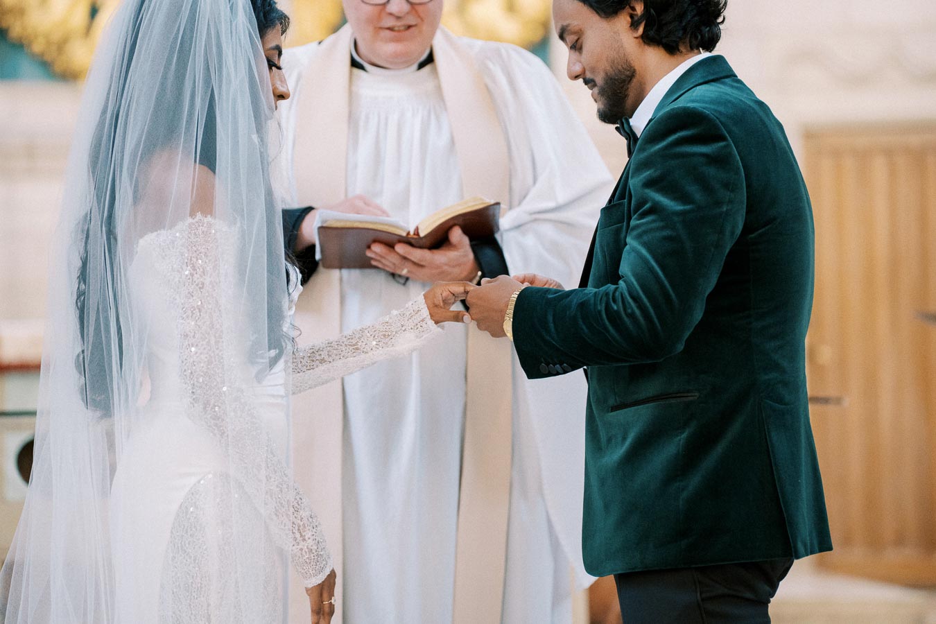 Wedding ceremony with bride in a white dress and veil holding hands with groom in a dark green suit, exchanging rings in front of a priest holding an open book.