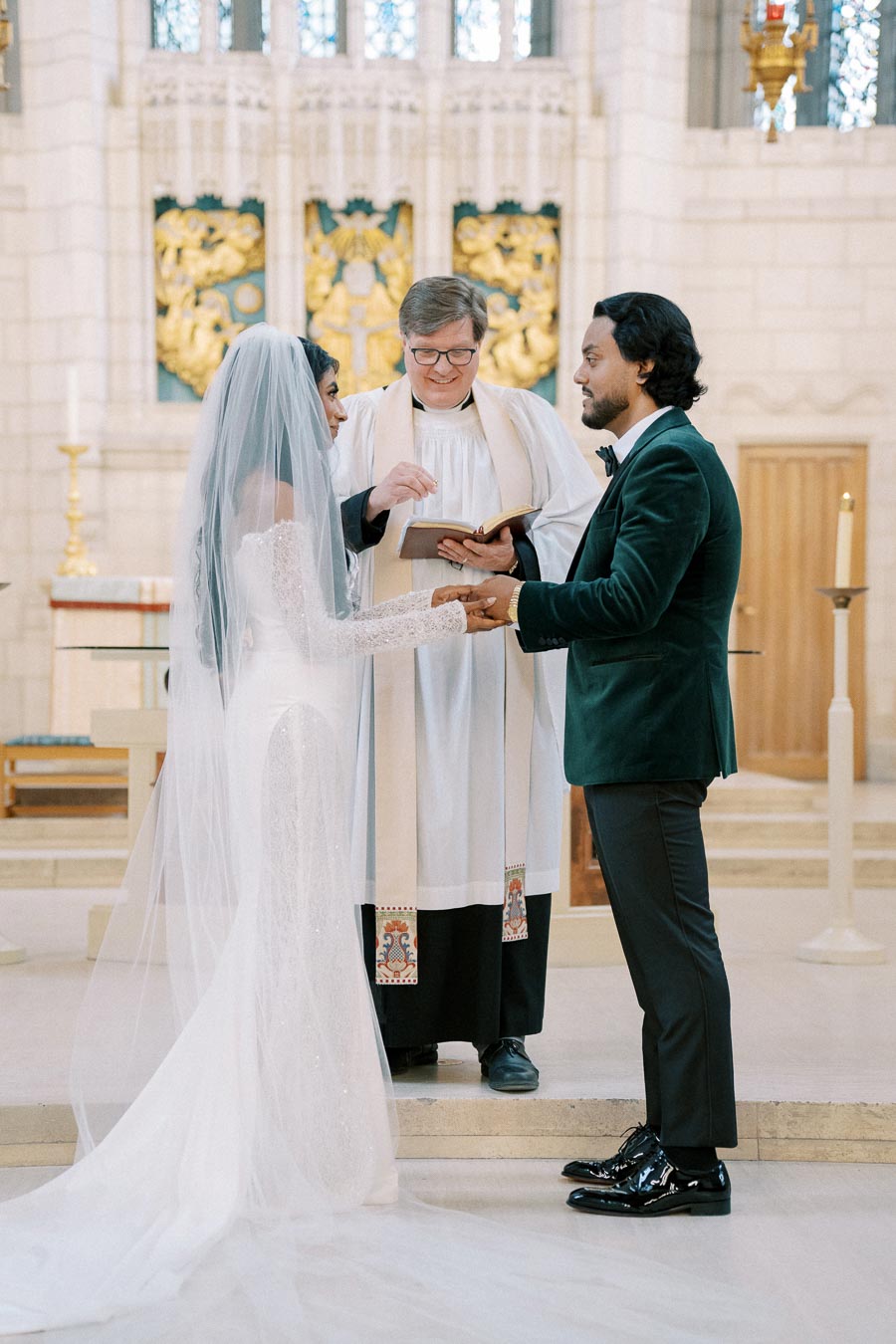 A bride in a white wedding gown and veil holds hands with a groom in a green suit at a church altar, officiated by a pastor during their wedding ceremony.