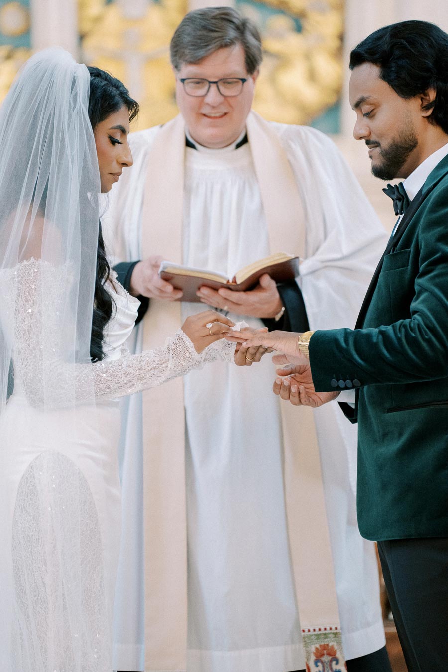 Bride and groom exchanging rings during a wedding ceremony officiated by a priest, bride in a white lace dress and veil, and groom in a dark suit.