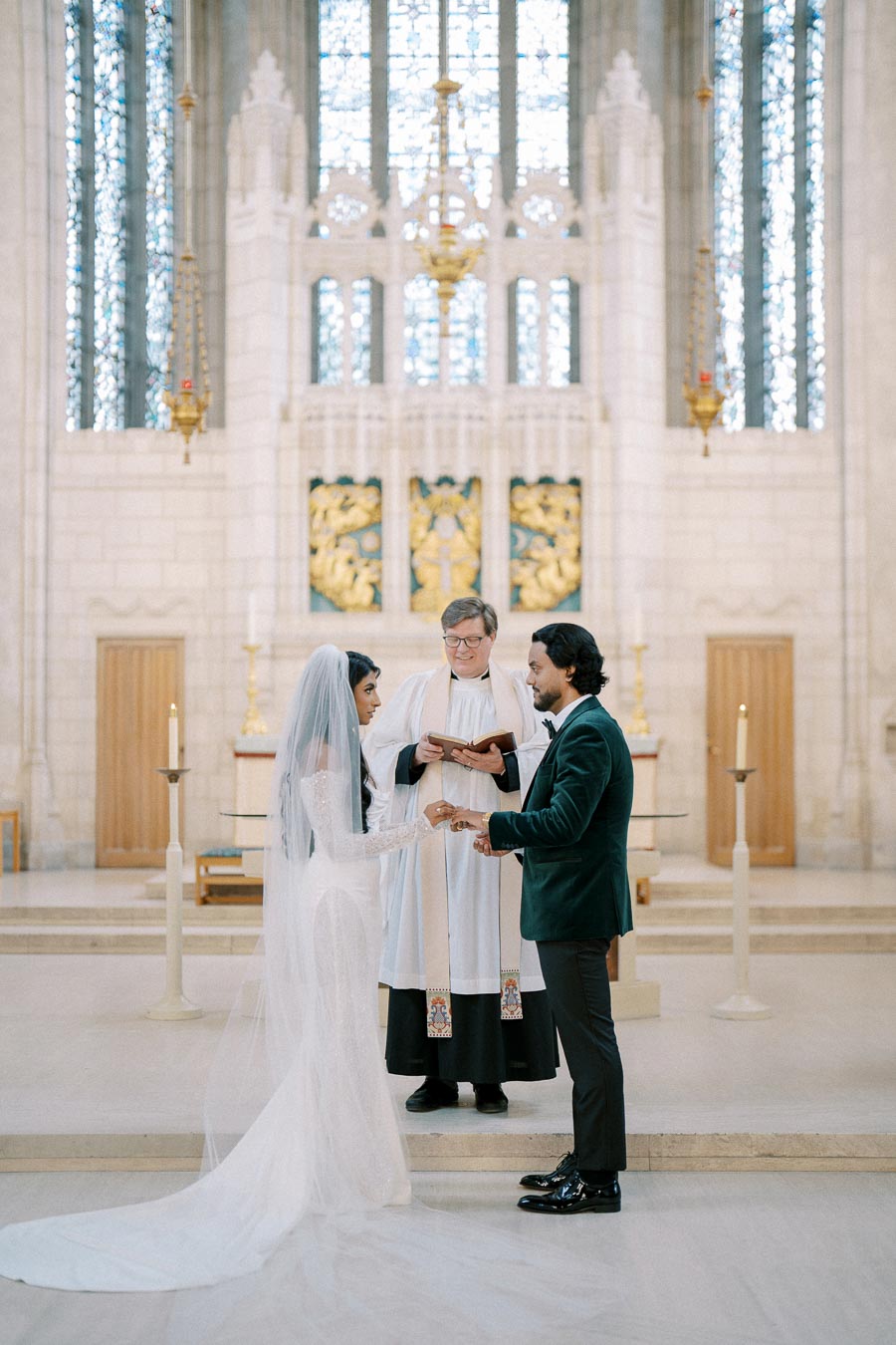 Bride and groom exchanging rings during a church wedding ceremony with a priest officiating, in an ornate, light-filled chapel.