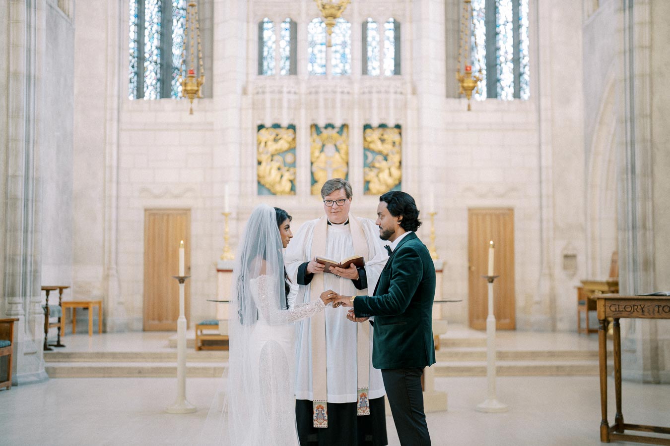 A bride and groom exchanging vows during their wedding ceremony in an ornate church, with an officiant overseeing the service. The bride is wearing a white gown and veil, and the groom is dressed in a dark suit. The church features elegant architecture and decor, including stained glass windows and candles.