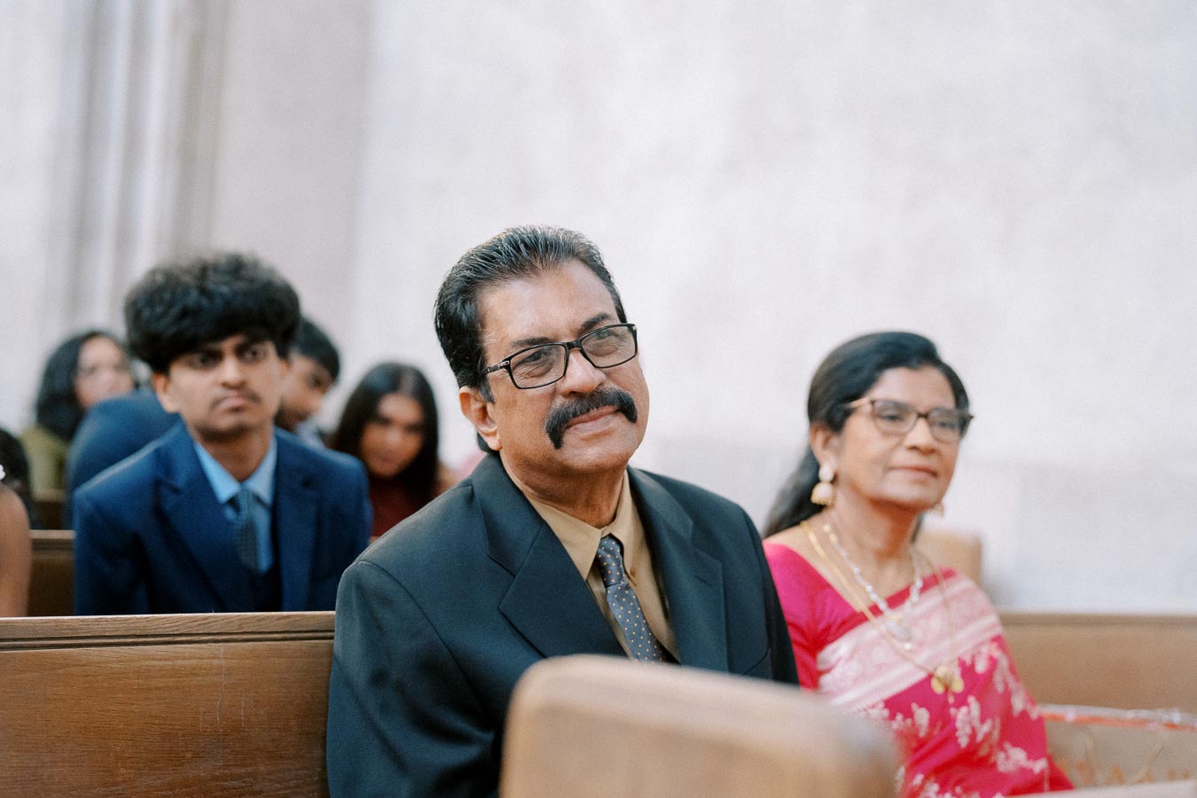 Group of people attending an indoor event, dressed in formal attire, with focus on a man in a black suit and woman in a pink traditional dress, seated in pews.
