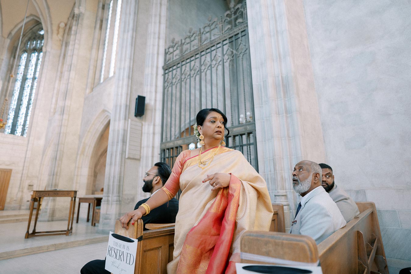A woman in a traditional sari standing in a church pew, surrounded by men in formal attire, under the high arches of a decorative church interior.