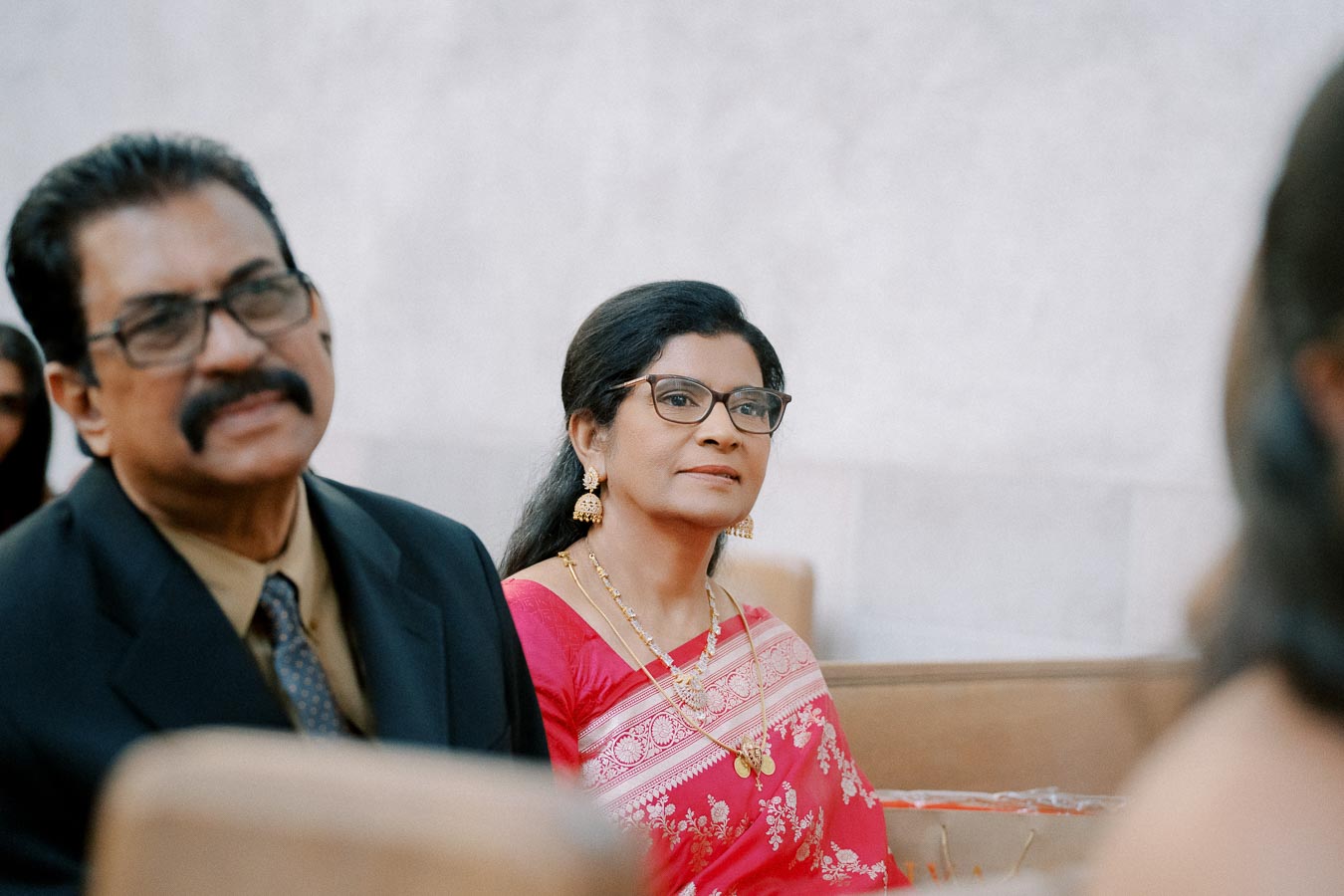 Two people in formal attire attending an indoor event, with a woman wearing a pink saree and gold jewelry, and a man in a suit and tie, both seated and looking ahead attentively.