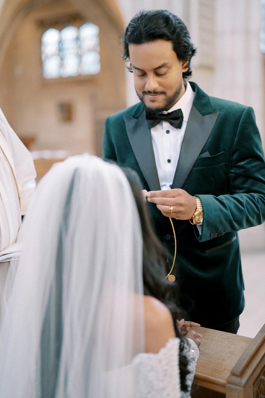 Groom in a velvet green tuxedo holds a ring during a wedding ceremony in a church, with the bride in a white dress and veil kneeling before him.