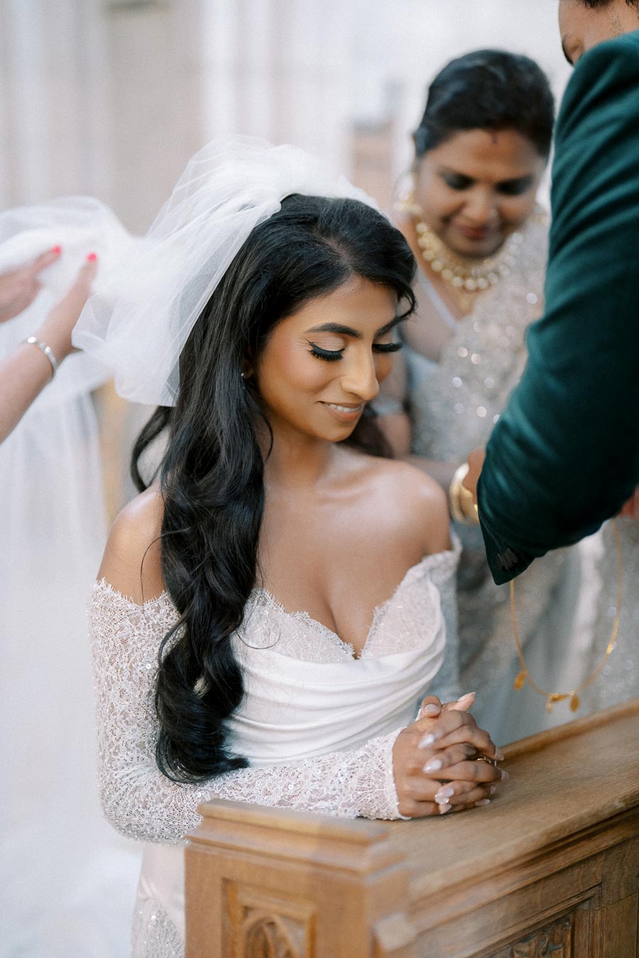 A bride in an elegant white lace off-shoulder dress and long black hair sits at a wooden kneeler, surrounded by family members, during a wedding ceremony.