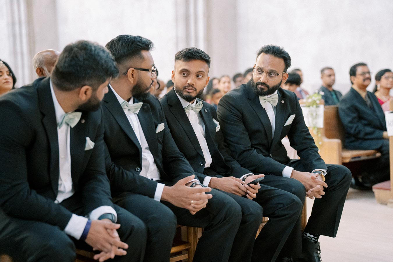 Four men in suits and bow ties seated at a wedding ceremony, engaged in conversation and surrounded by an audience.