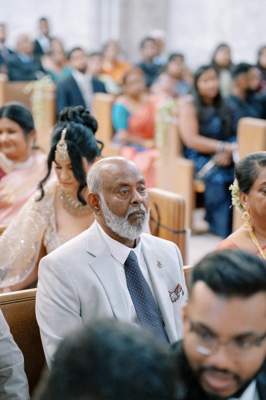 Elderly man wearing a light grey suit attending a formal event in a crowded room filled with seated guests.