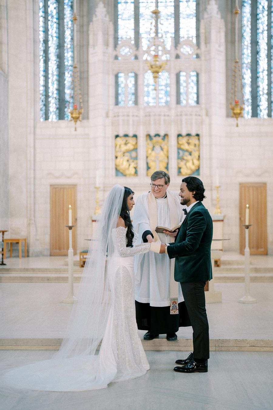 A bride and groom exchange vows in a beautiful church ceremony, officiated by a priest wearing a white robe. The bride wears a sparkling white gown and veil, while the groom is in a dark suit, set against an ornate altar backdrop with stained glass windows.