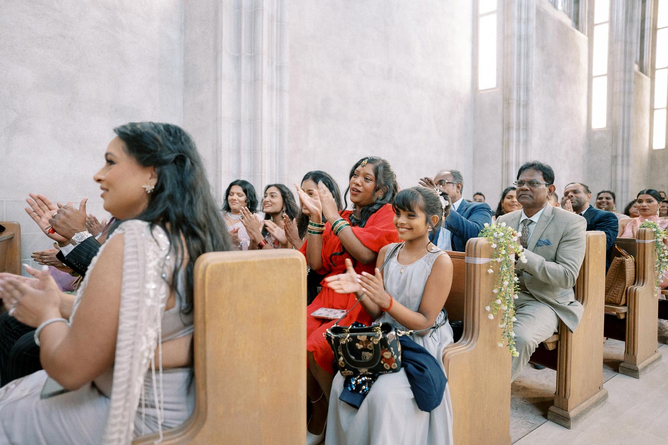 Guests seated in a chapel, clapping and smiling during a wedding ceremony, with wooden benches adorned with floral decorations and natural light streaming through tall windows.