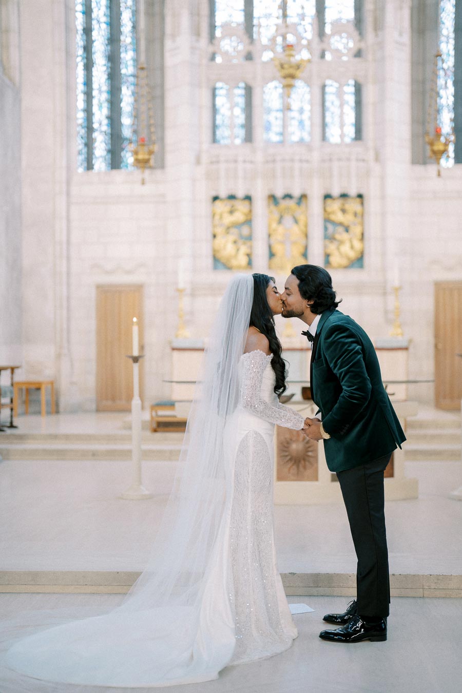 A bride and groom share a kiss inside a beautifully decorated church, with the bride wearing a white wedding dress and veil, and the groom in a dark suit, standing in front of a stained glass window and altar.