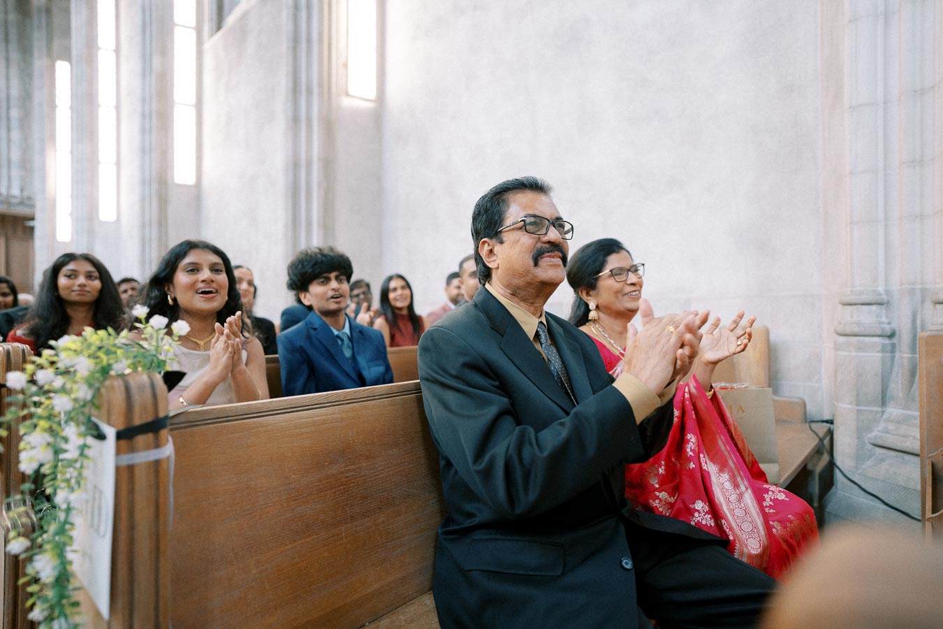 Audience clapping in a church setting during a formal event, featuring diverse individuals in traditional and modern attire seated on wooden pews, surrounded by floral decorations.