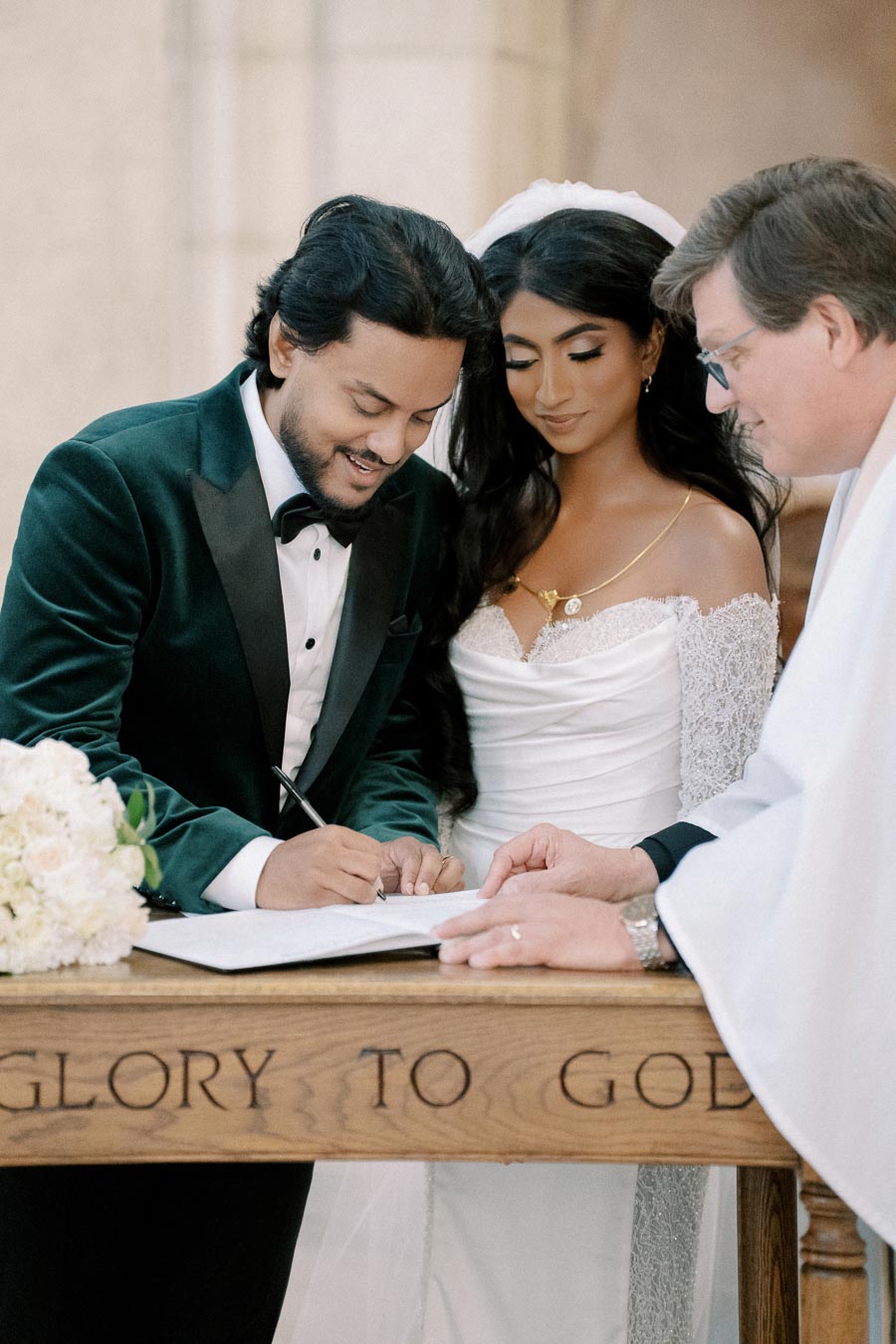 Wedding ceremony with couple signing marriage certificate next to officiant at altar, bride wearing elegant white gown and groom in green velvet suit.