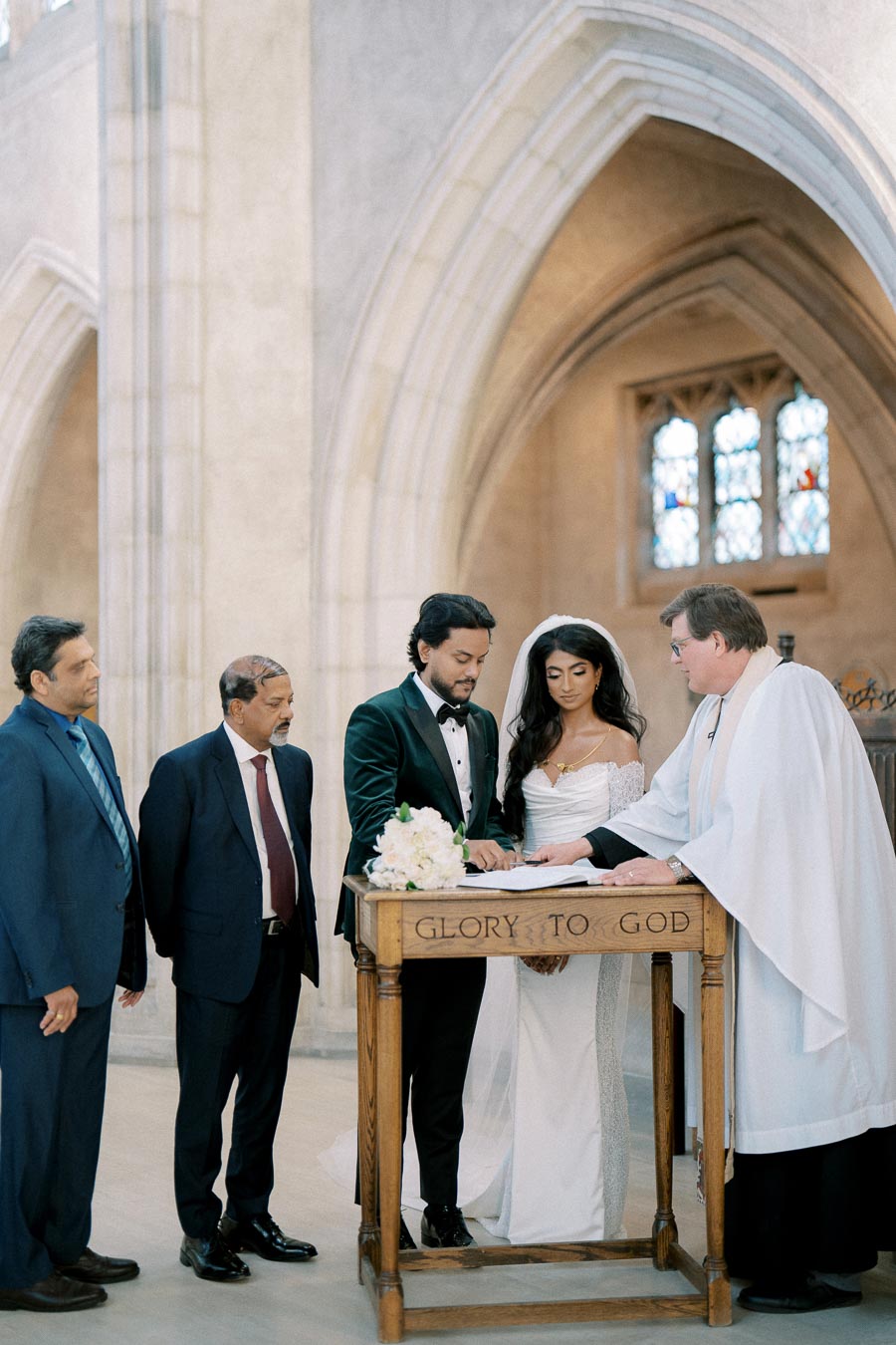 A wedding ceremony in a church with a couple signing the marriage certificate, surrounded by officiant and two witnesses, with stained glass windows in the background.