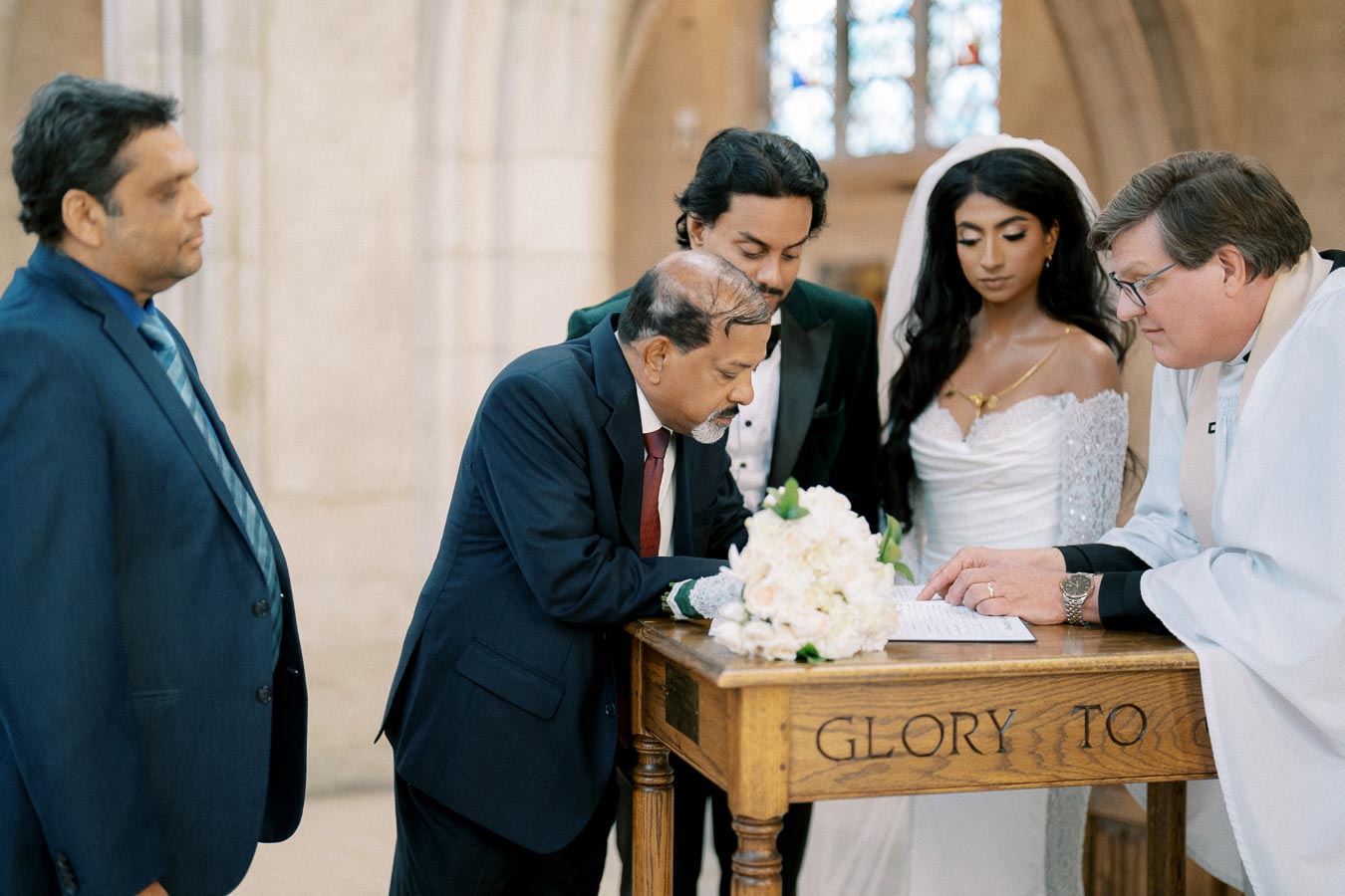 A bride and groom stand in a church beside an officiant and witnesses, all focused on a document atop a wooden table with flowers. The church setting is bright and historic, creating an elegant wedding atmosphere.