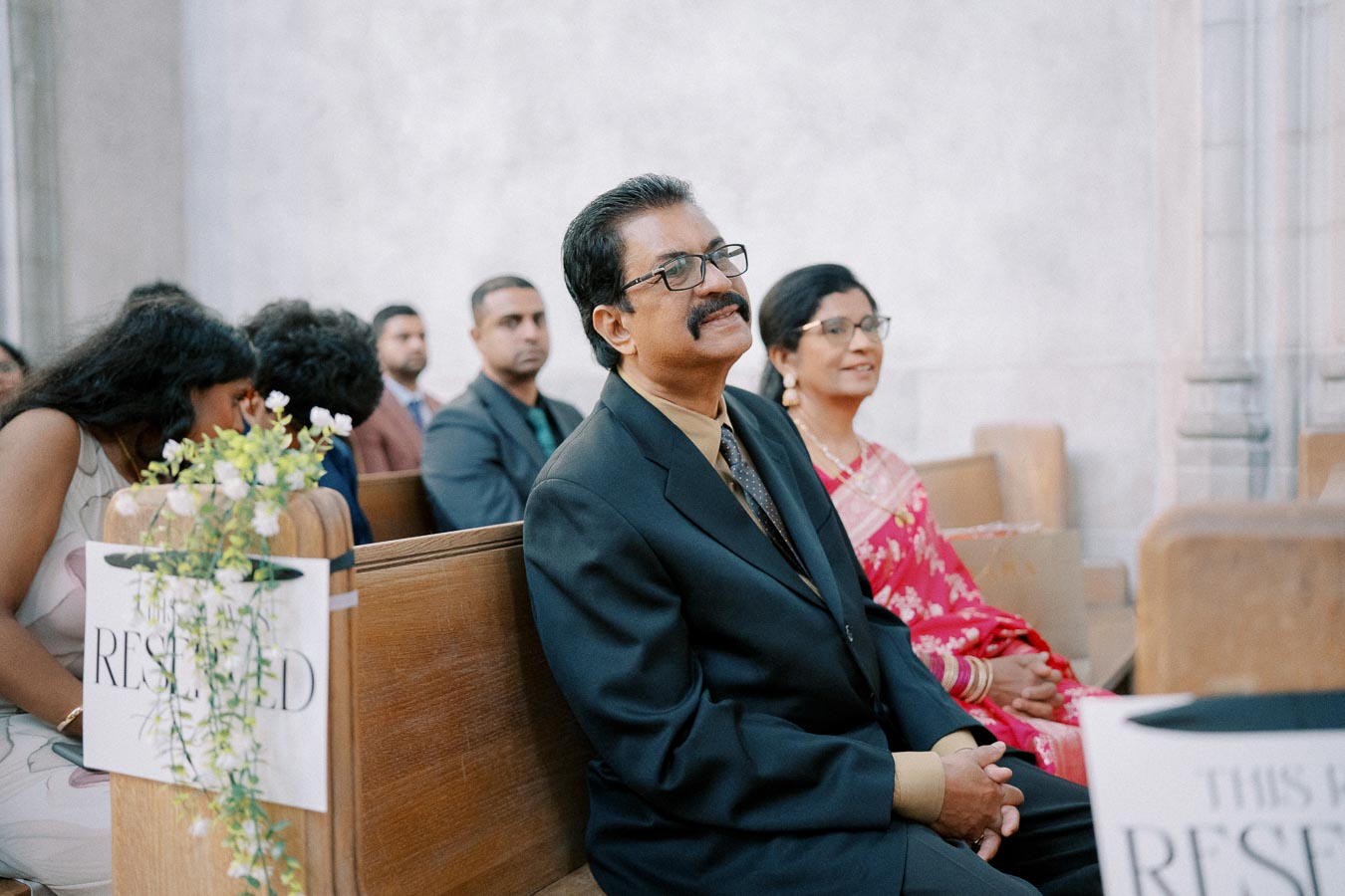 A middle-aged couple attentively seated in a church pew during a formal event. The man, wearing a dark suit and tie, and the woman in a red, floral-patterned sari are surrounded by other attendees. A “reserved” sign with white flowers is visible on the pew, indicating a special occasion, likely a wedding or ceremony.
