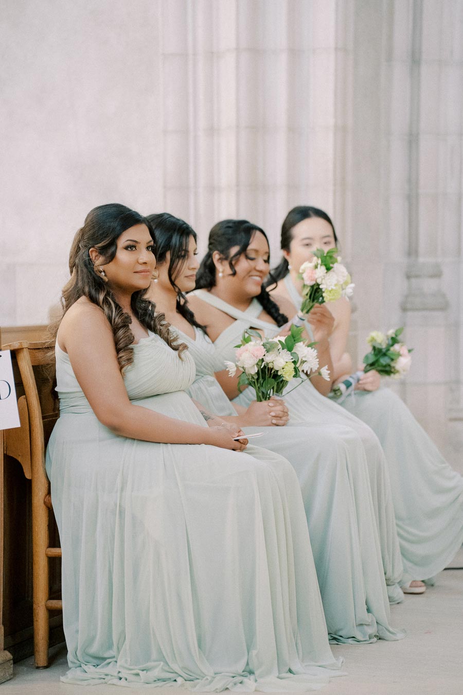 Bridesmaids seated in light green dresses holding bouquets, smiling and engaged in a wedding ceremony at an elegant venue.