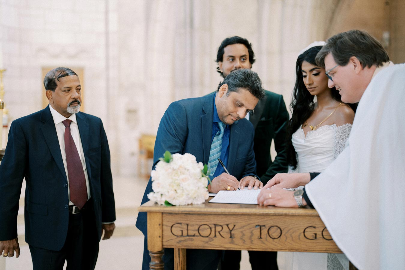 Wedding ceremony in a church with bride, groom, and officiant signing the marriage certificate on a wooden table with flowers, surrounded by guests.