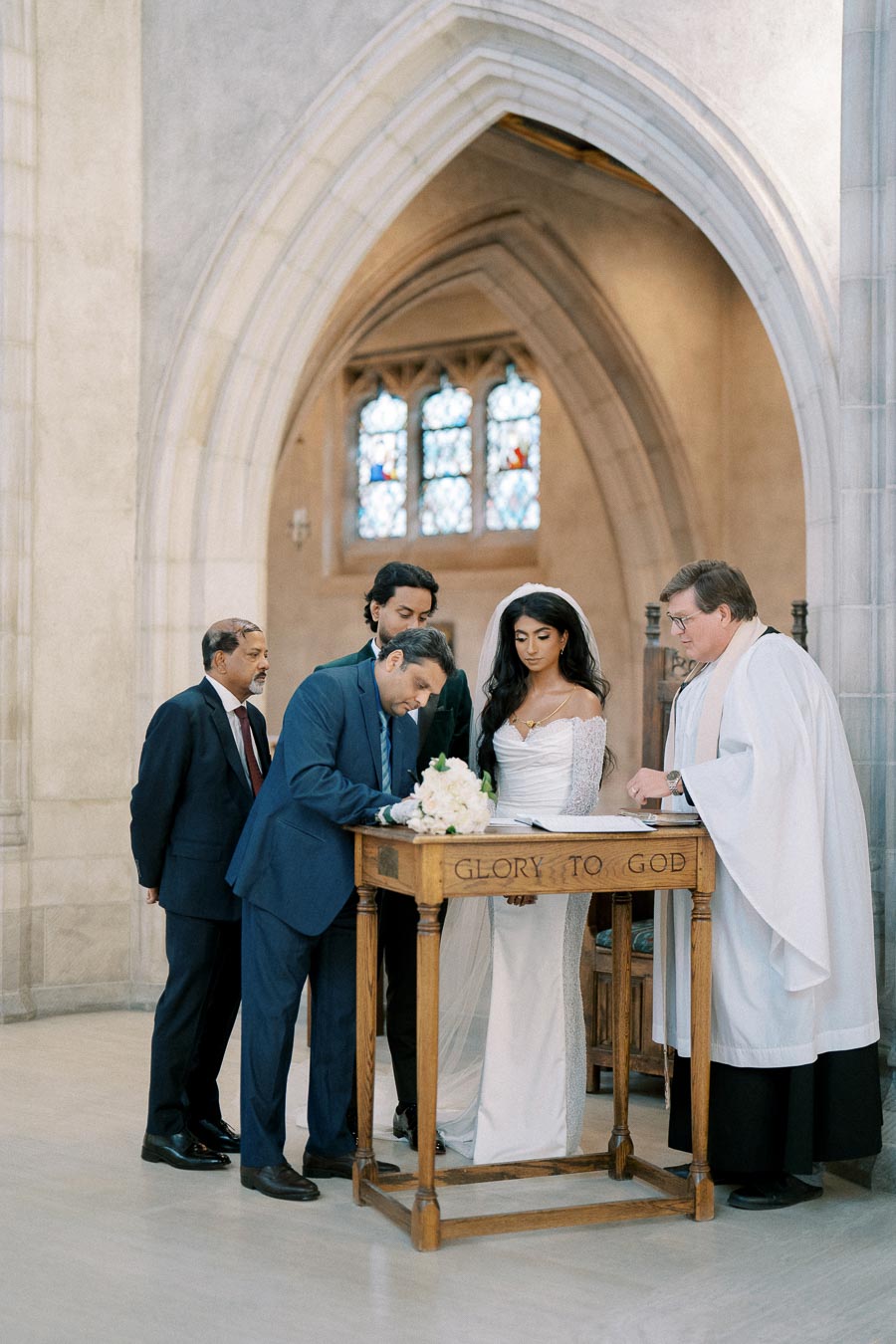A bride and groom sign their marriage certificate at a wooden table with Glory to God engraved on it inside a church, accompanied by a priest and two witnesses, with stained glass windows in the background.