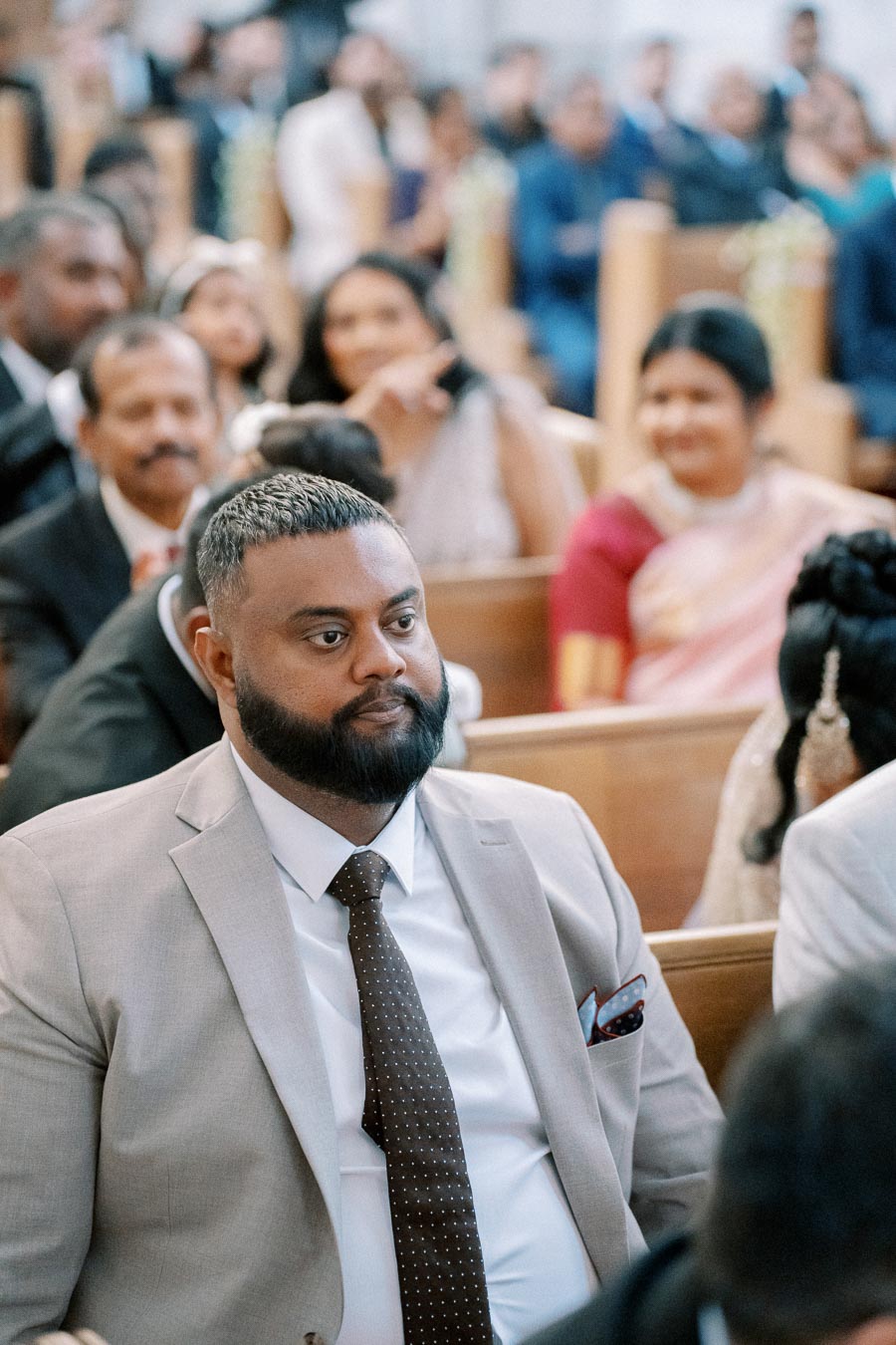 A man in a light gray suit and dotted tie sits attentively in a church pew among other formally dressed guests, suggesting a wedding or formal ceremony setting.