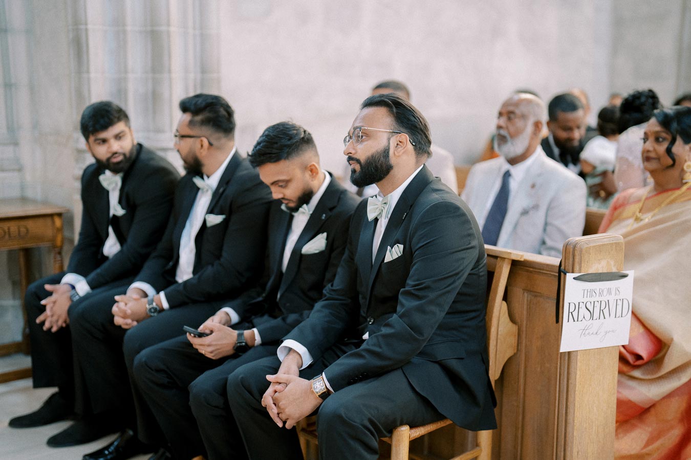 Formal wedding guests seated in a reserved church pew, with four men in black suits and bow ties attentively waiting.