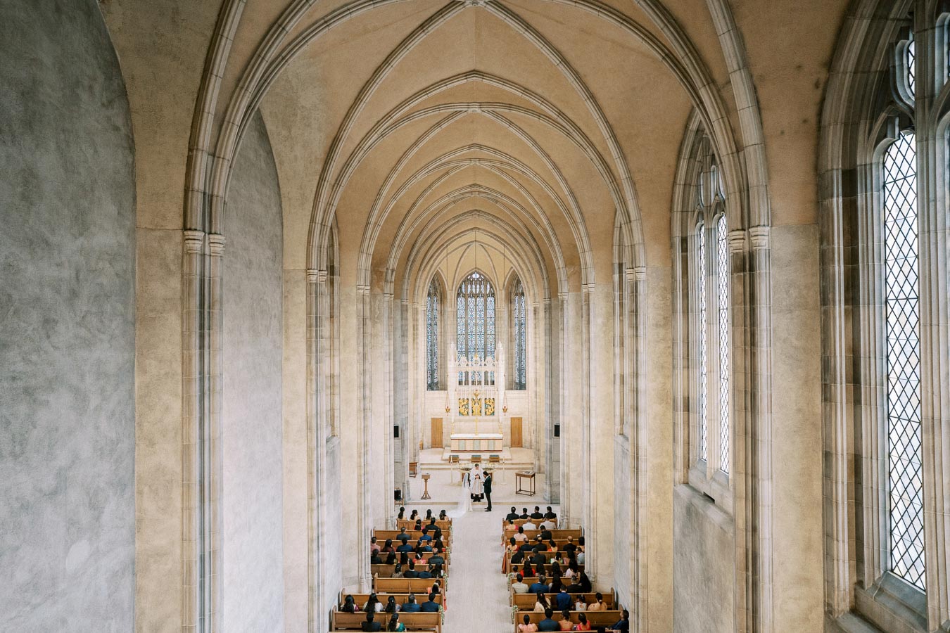 A breathtaking view of a grand cathedral interior with vaulted ceilings and stained glass windows; a wedding ceremony is taking place at the altar with guests seated in wooden pews.