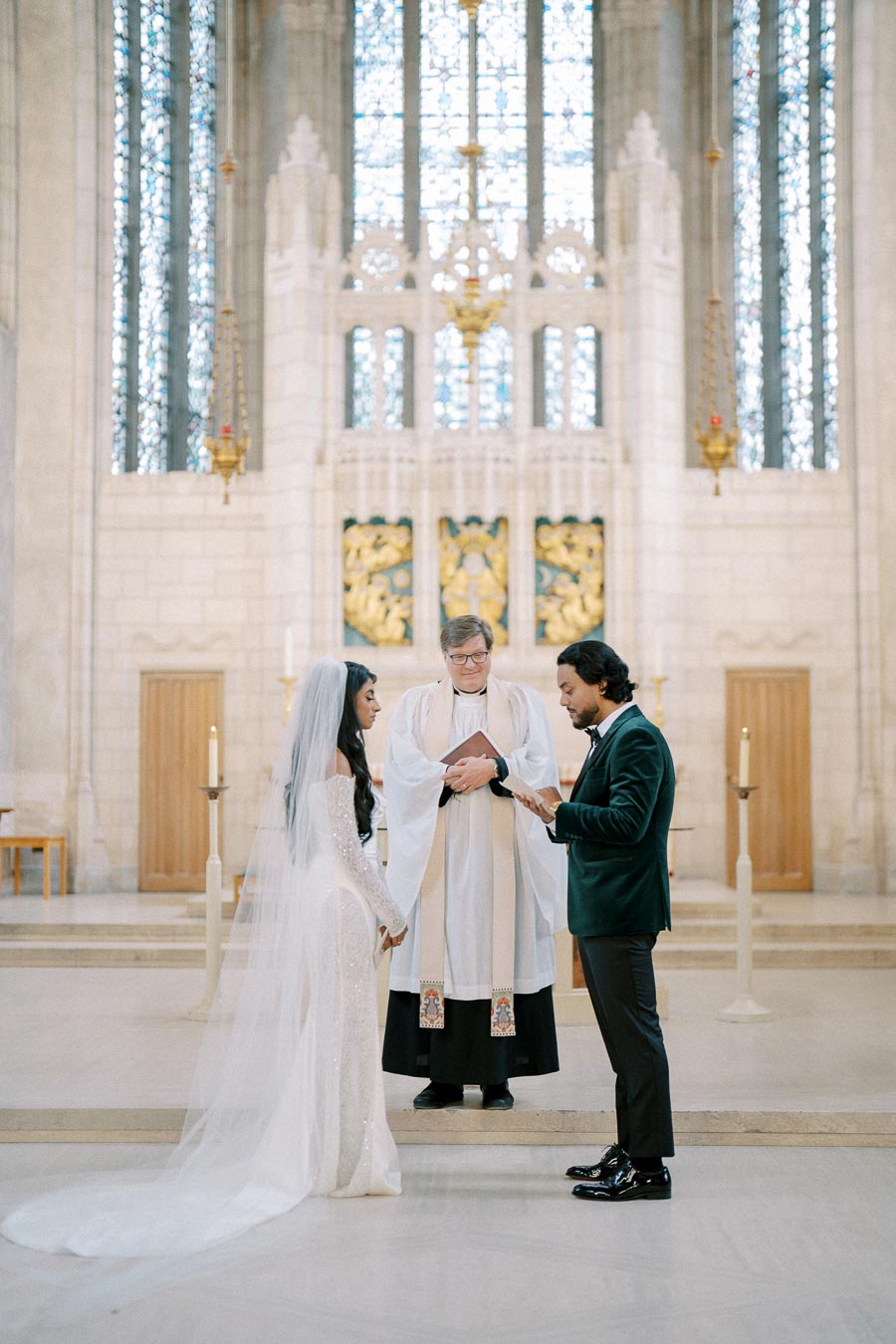 A bride in an elegant white gown with a long veil and a groom in a dark suit exchanging vows in a beautiful church setting, accompanied by a priest. Stained glass windows and candles create a serene and solemn atmosphere.