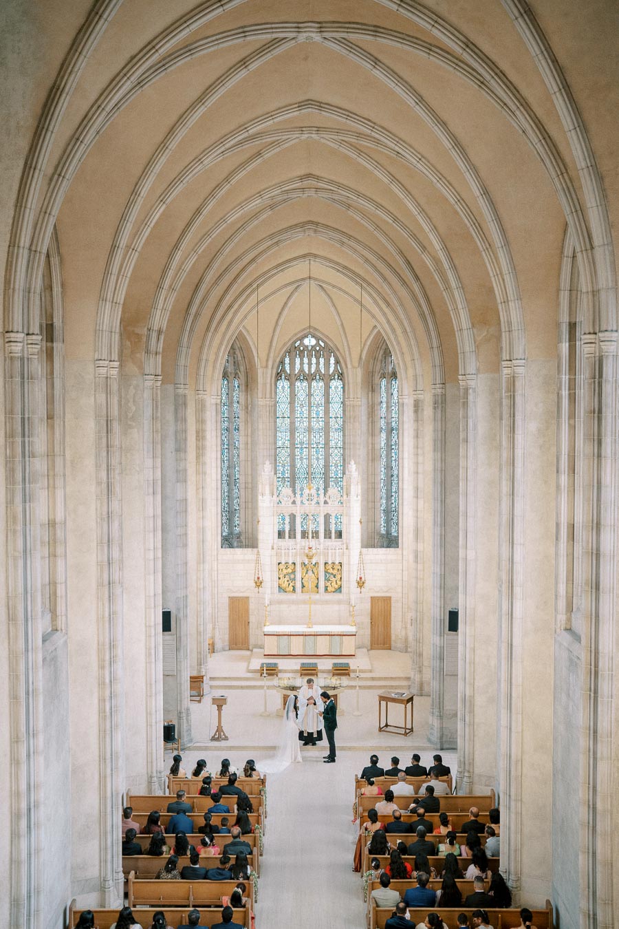 Wedding ceremony in a grand cathedral with high vaulted ceilings, ornate stained glass windows, and guests seated in pews.