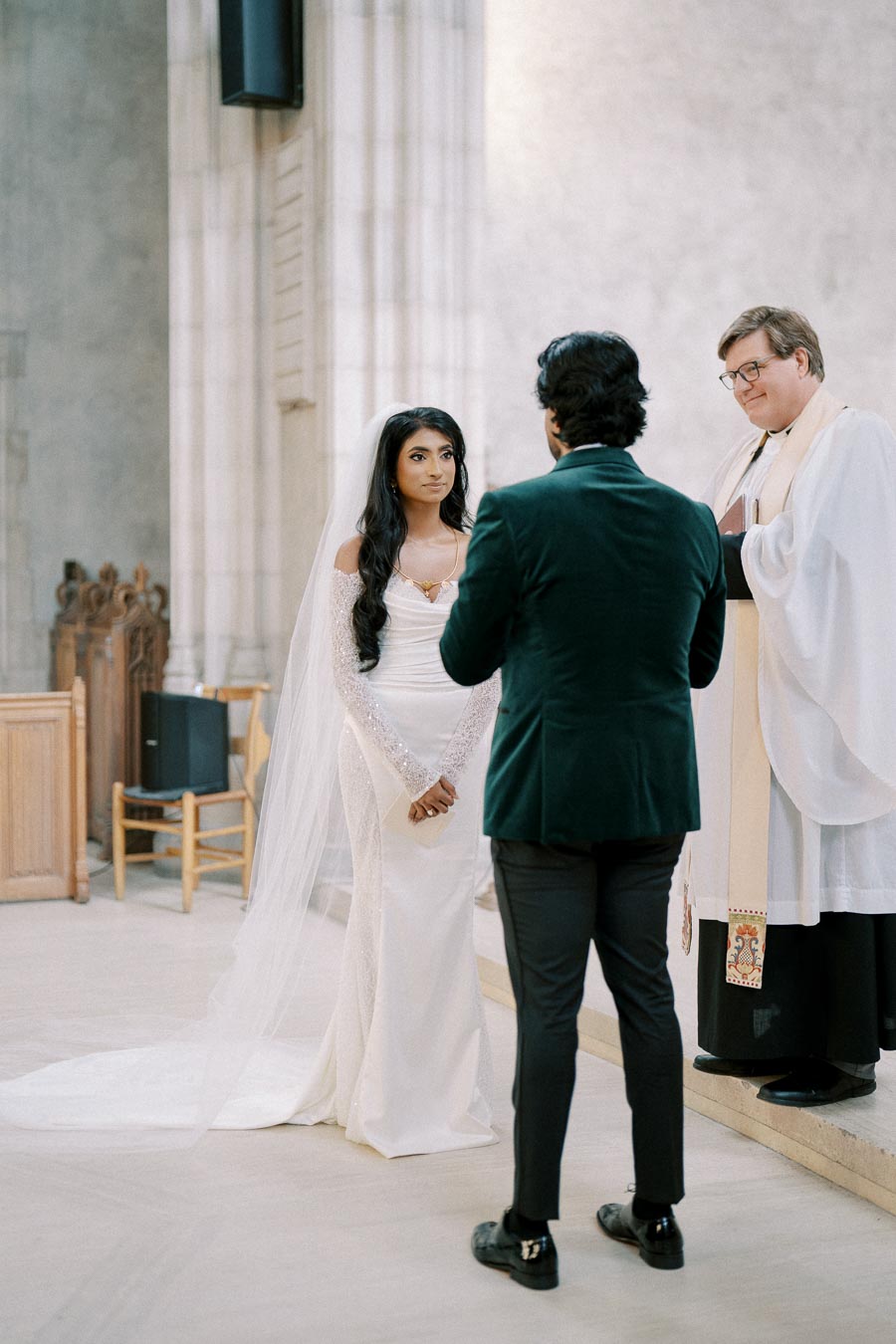 Bride and groom standing before officiant during wedding ceremony inside a church, bride in elegant white gown with long veil, groom in dark suit.