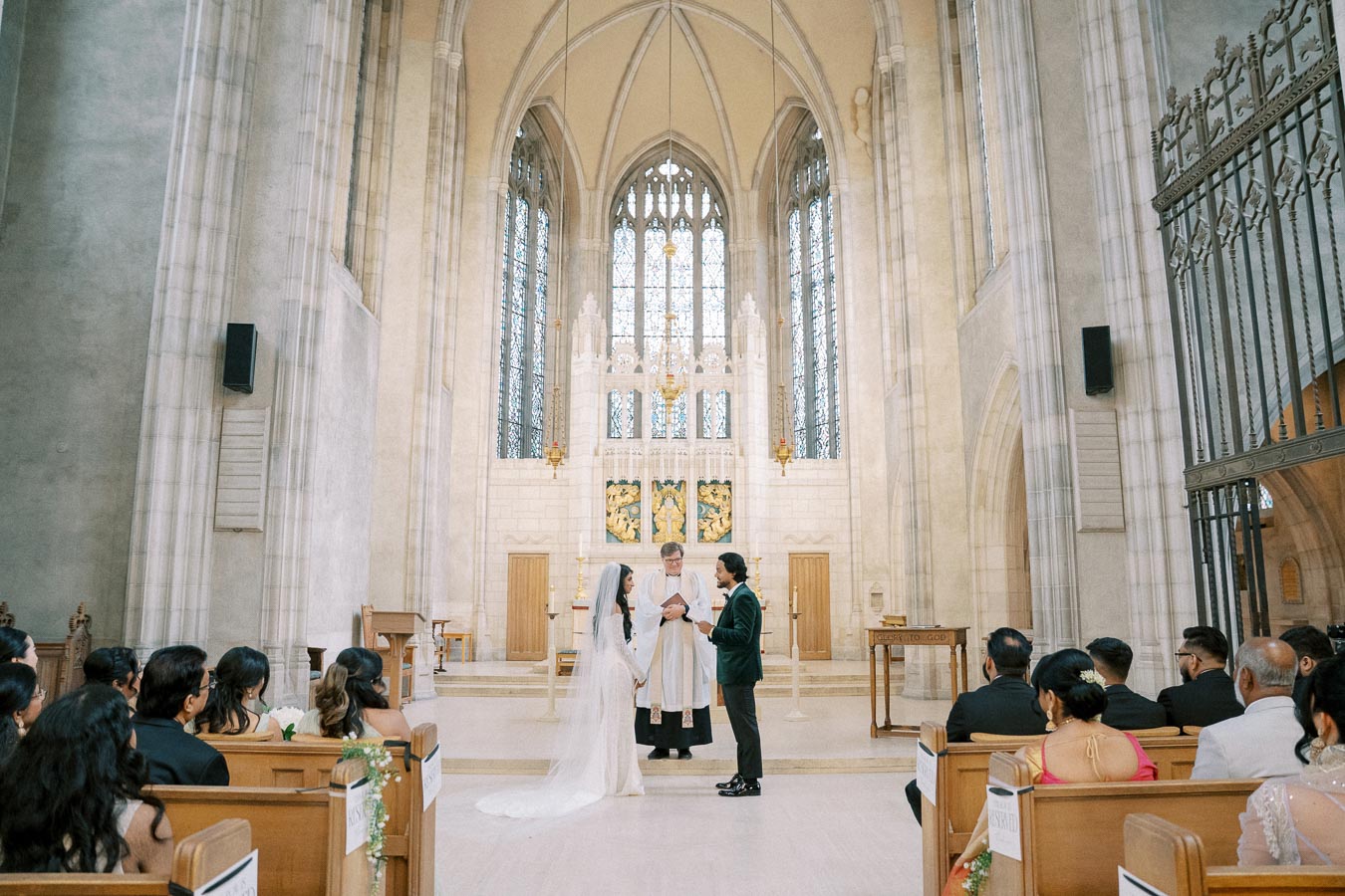 Wedding ceremony in a grand cathedral, featuring a couple exchanging vows at the altar with a priest. The elegant architecture includes arched ceilings and stained glass windows. Guests are seated, observing the intimate moment.