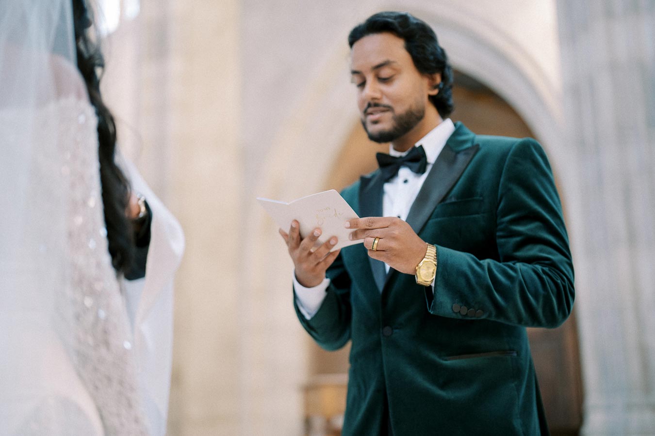 A groom in a velvet tuxedo reads vows during a wedding ceremony inside a cathedral.