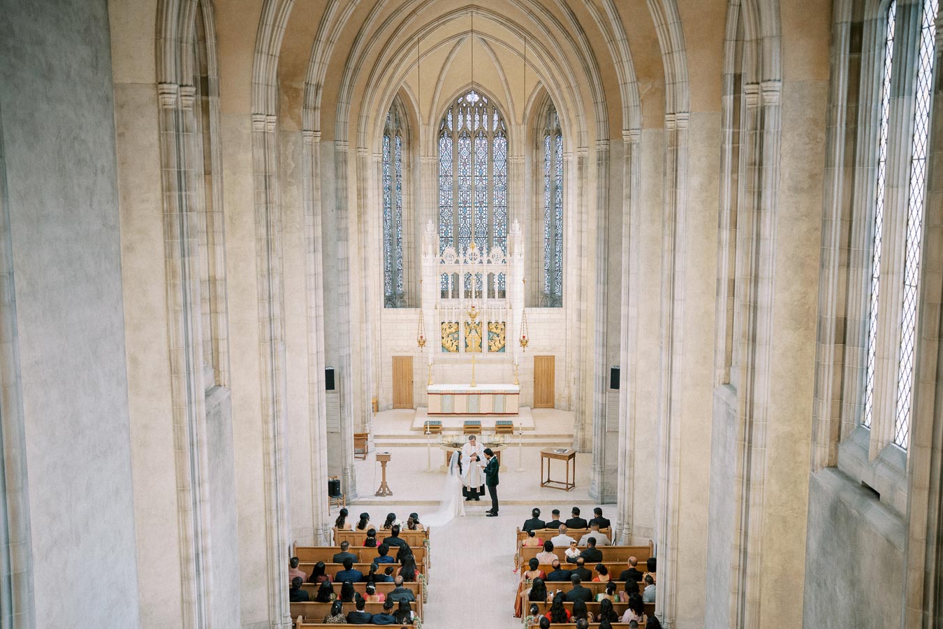 Cathedral interior with high ceilings hosting a wedding ceremony, featuring stained glass windows and guests seated in pews.