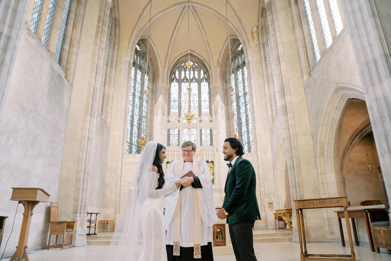 A bride and groom stand before a priest during their wedding ceremony in a grand cathedral, featuring stained glass windows and high vaulted ceilings.