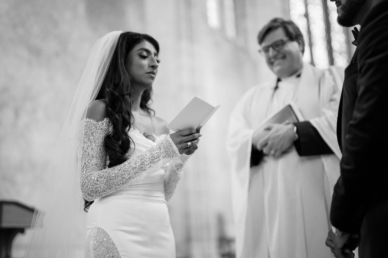 Black and white image of a bride reading vows during a wedding ceremony, standing with a priest and groom in a church setting.