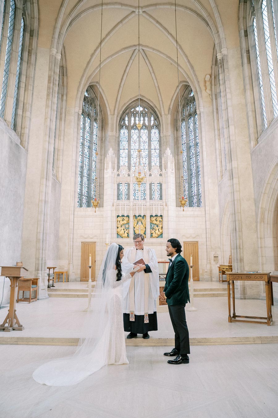 Couple exchanging vows in a grand church setting with ornate stained glass windows and high arches, officiated by a clergyman.