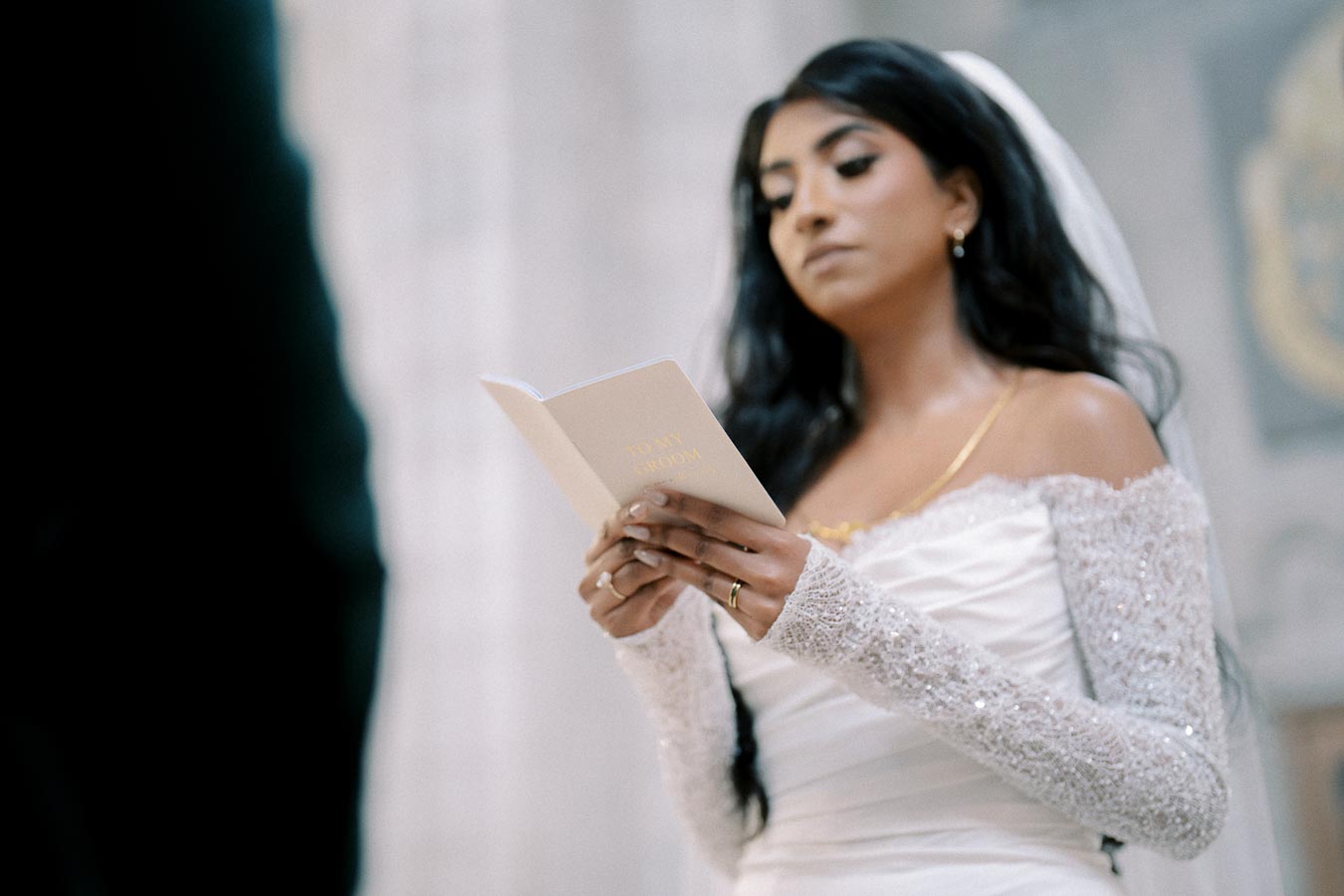 Bride reading vows during wedding ceremony, wearing a white lace wedding dress and veil, holding a booklet titled To My Groom.