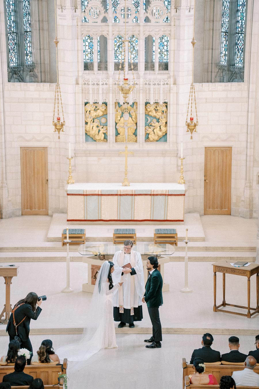 A bride and groom exchange vows at the altar of a beautifully decorated church with a priest officiating the wedding ceremony, surrounded by family and friends, captured by a photographer.