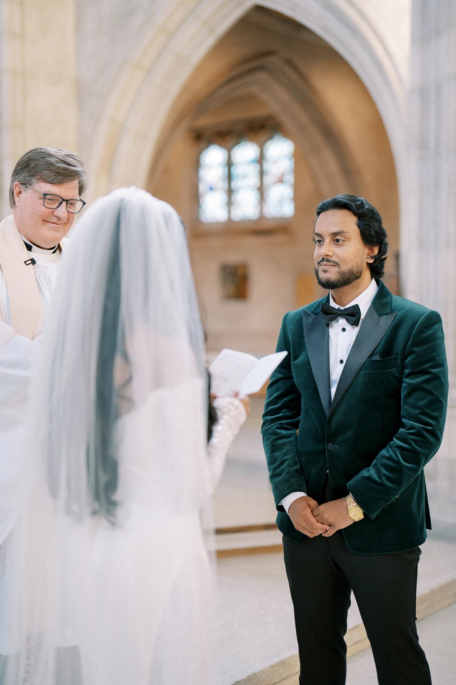 Bride and groom exchanging vows during a wedding ceremony in a church, with the groom wearing a green velvet suit and a priest officiating. The bride is holding wedding vows in her hand.